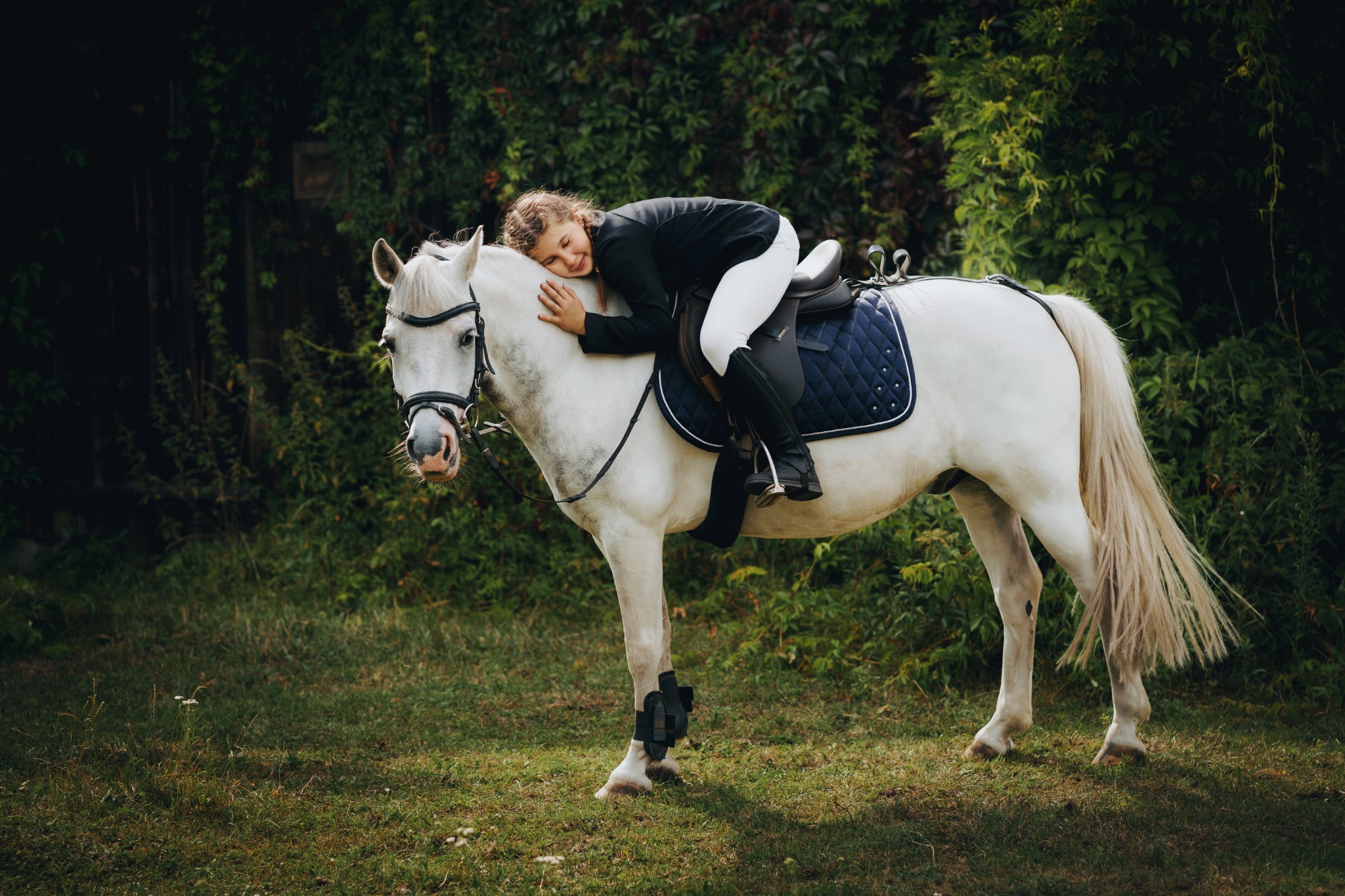 Młoda dziewczyna w stroju jeździeckim, oparta o białego konia z zamkniętymi oczami w zalesionym otoczeniu.