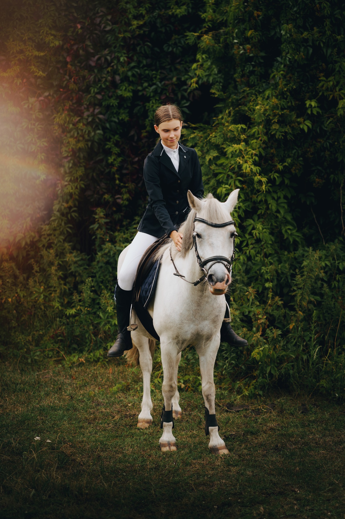 Młoda kobieta w stroju jeździeckim siedzi na białym koniu na tle zieleni.