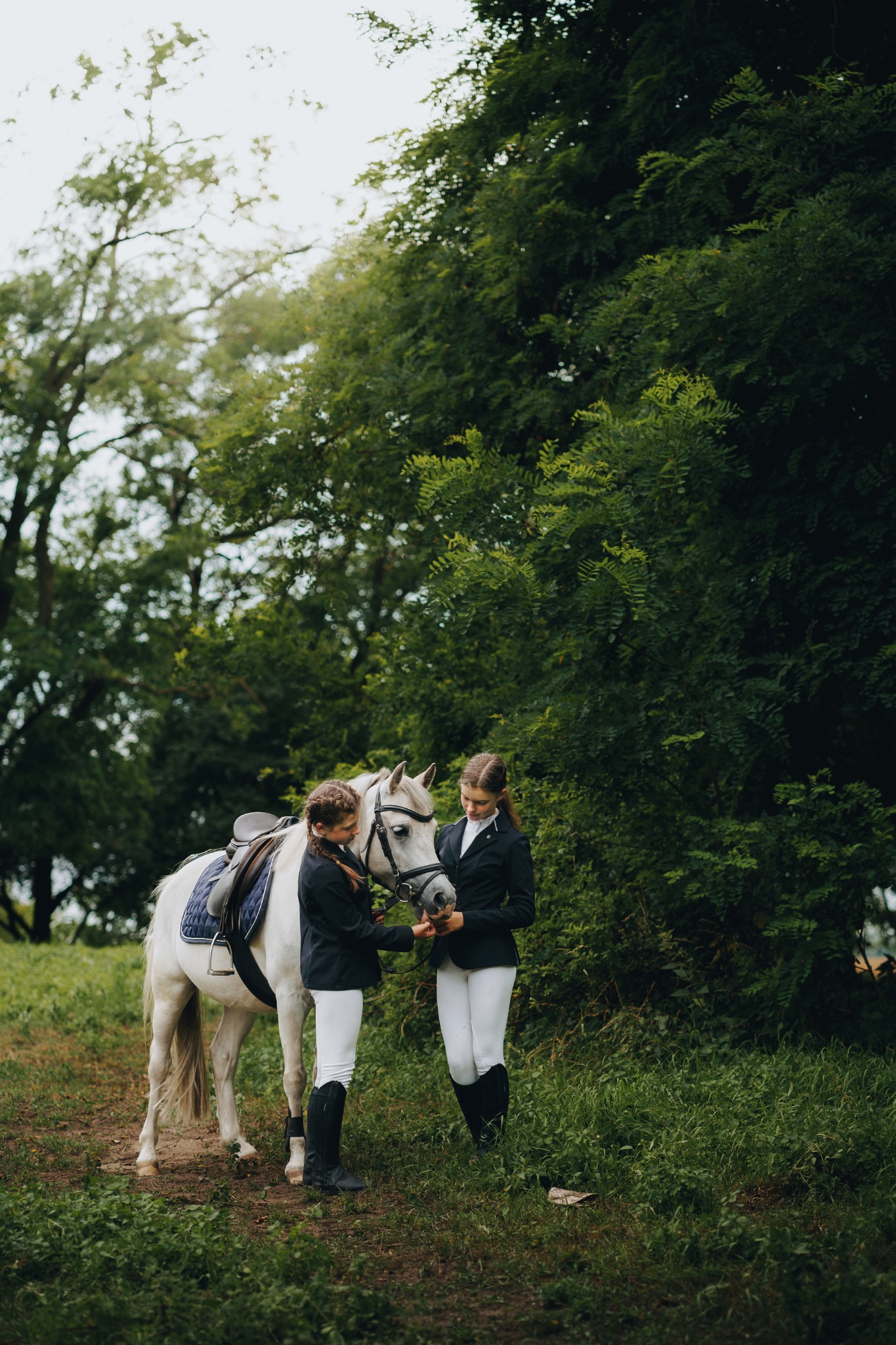 Dwie młode kobiety w strojach jeździeckich głaszczą białego konia z siodłem na tle zieleni.