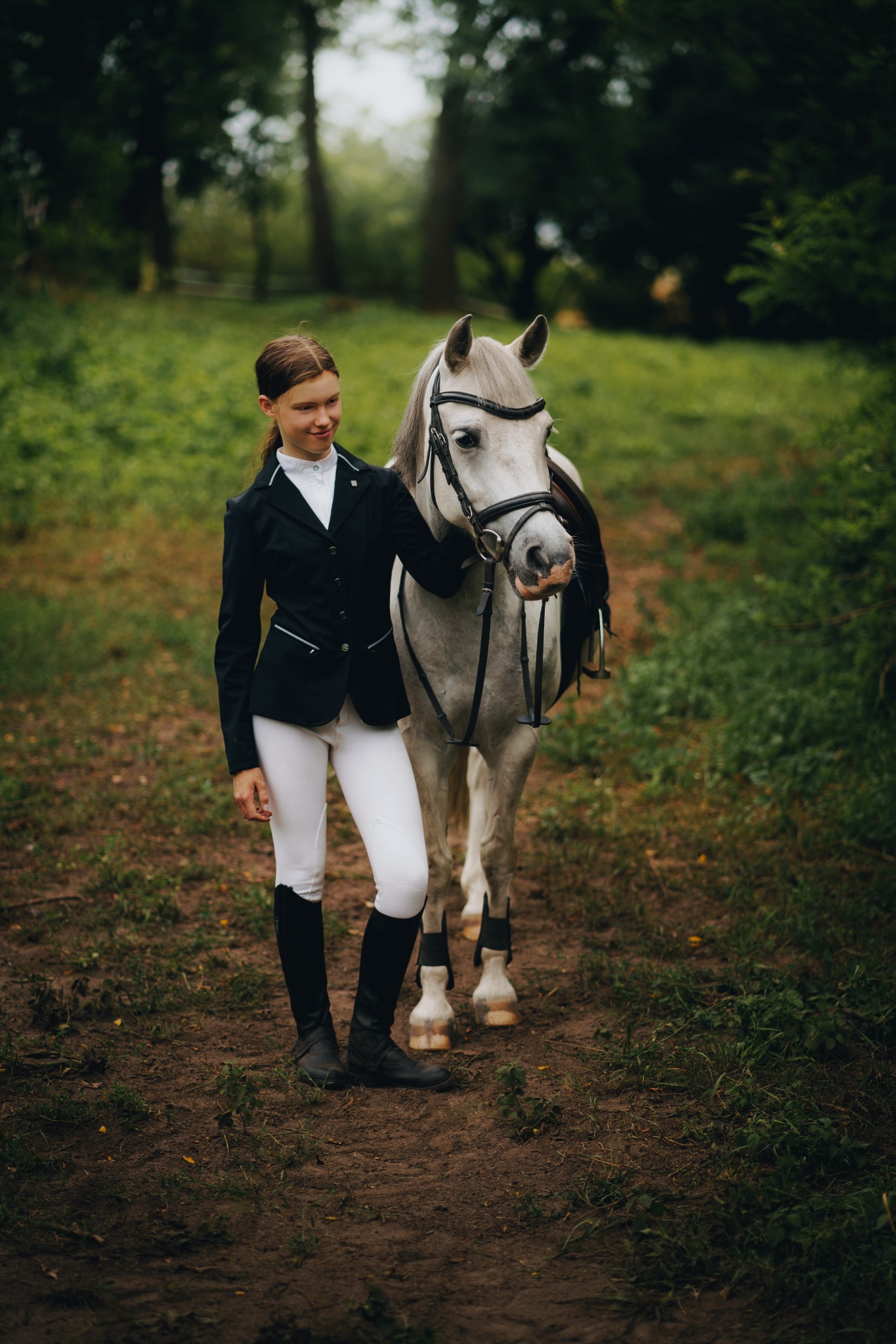 Młoda kobieta w stroju jeździeckim, czarnej marynarce i białych bryczesach, stoi obok siwego konia w uprzęży na leśnej ścieżce.