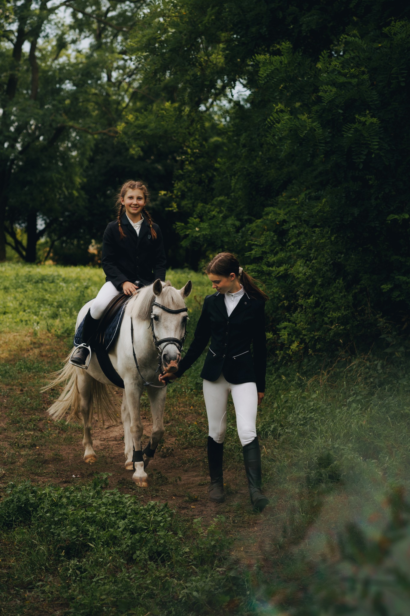 Dziewczynka w czarnej kurtce jeździeckiej na białym koniu prowadzonym przez kobietę w białych bryczesach na tle zieleni.