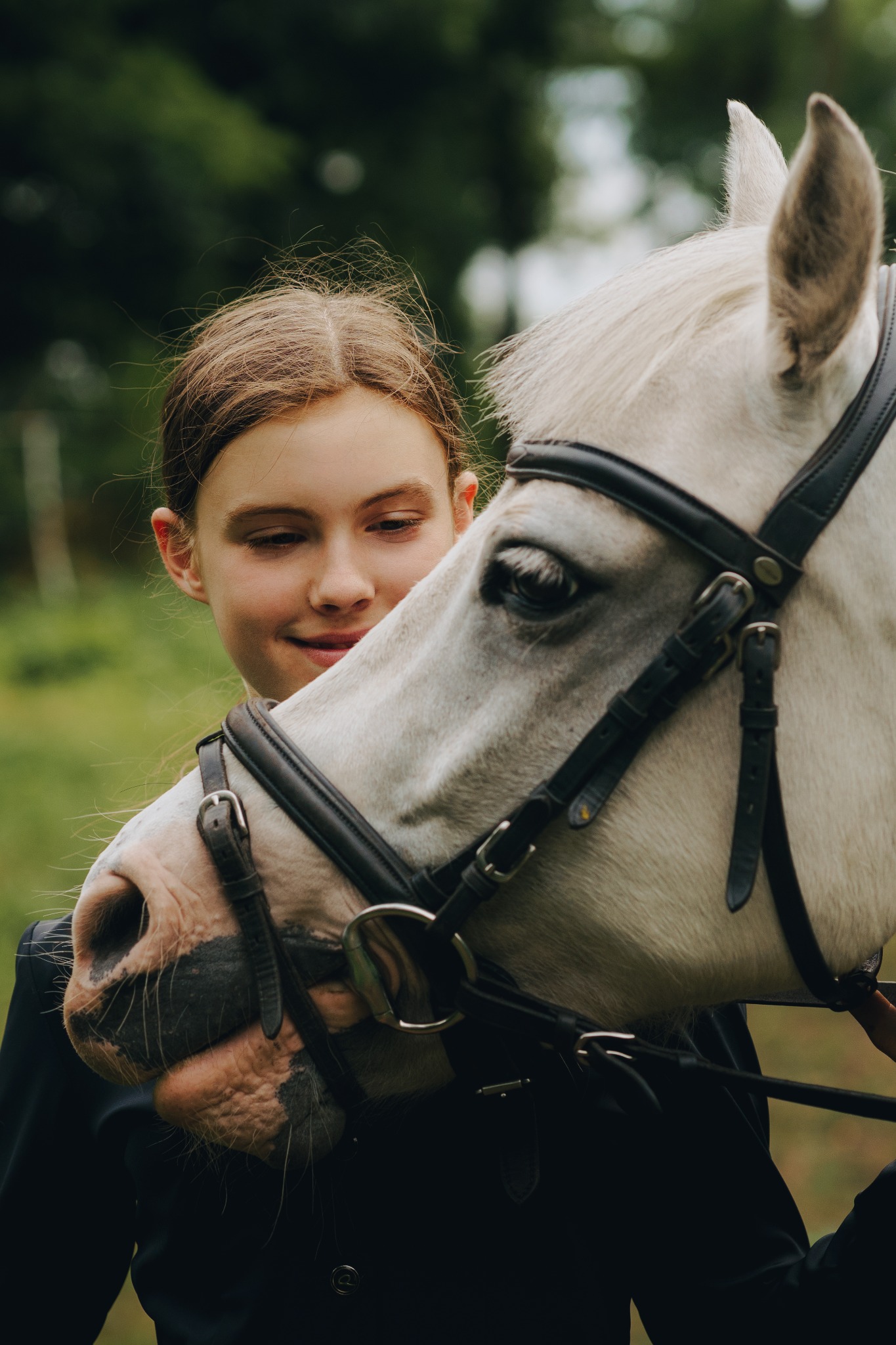 Młoda dziewczyna w czarnym stroju obejmuje białego konia w czarnym ogłowiu na tle zieleni.