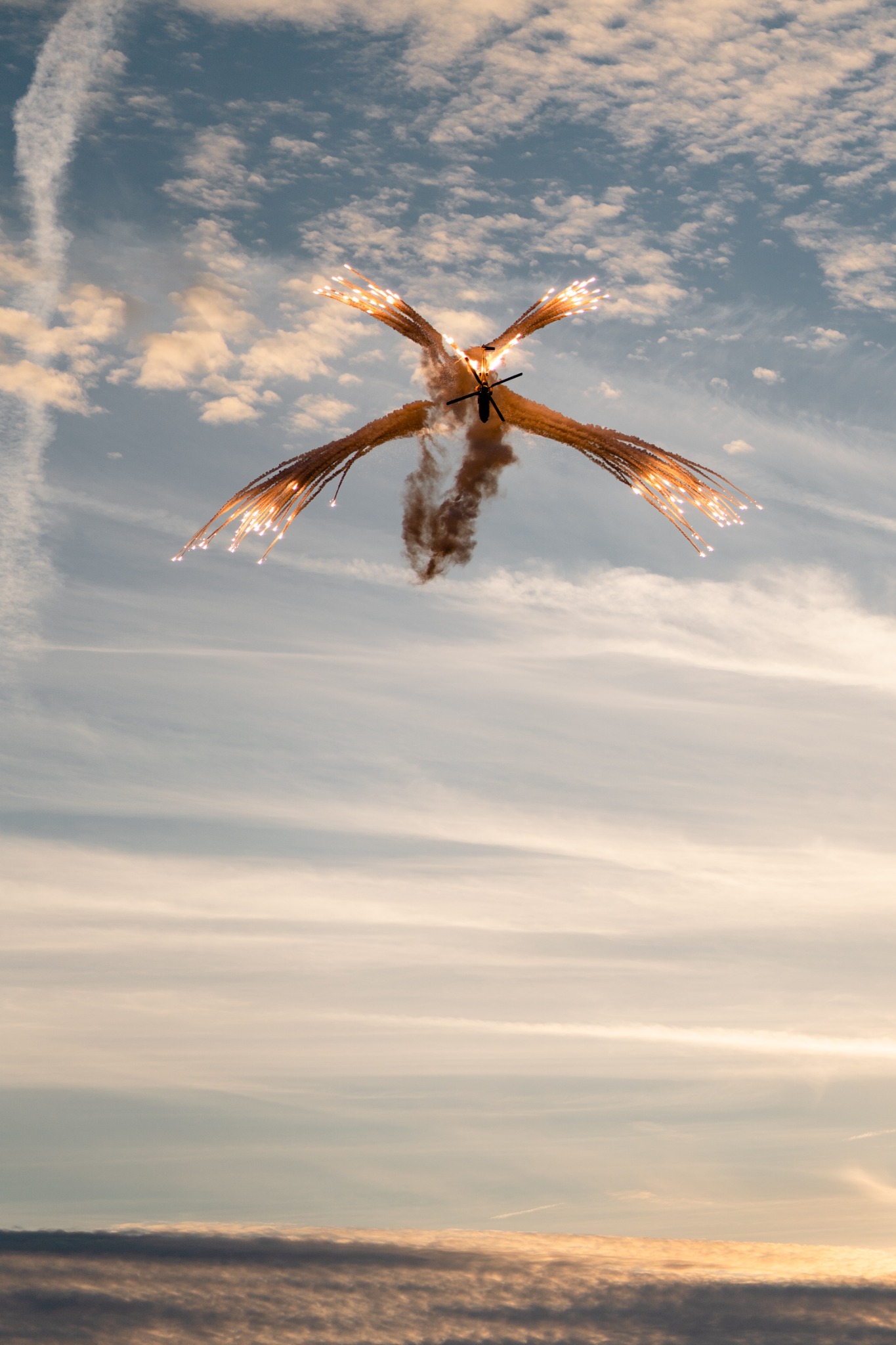Helikopter w locie na tle nieba, wystrzeliwujący flary ochronne, tworzące efektowne promienie światła.