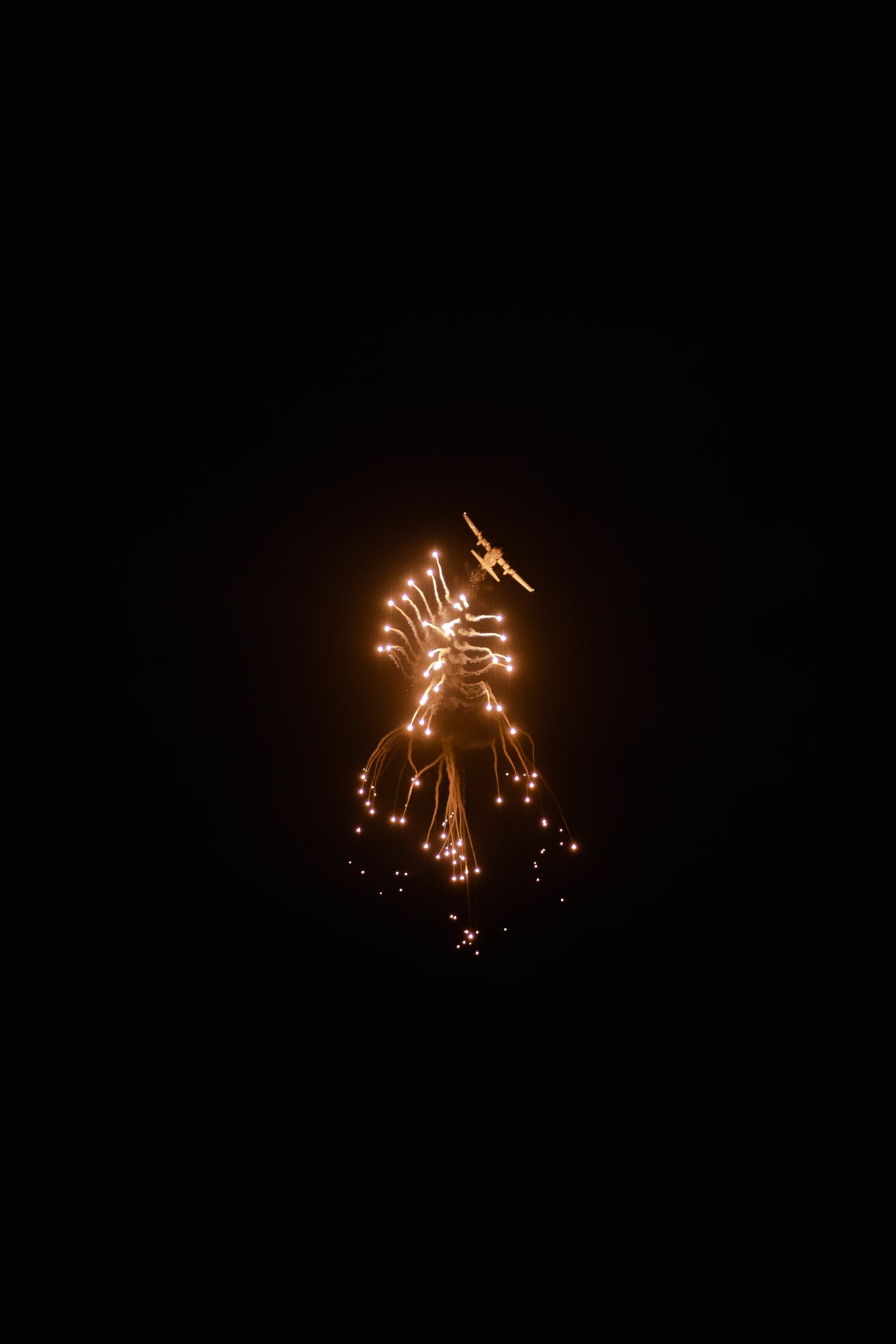 Nocne niebo rozświetlone złotymi fajerwerkami, z sylwetką samolotu widoczną na tle eksplozji.