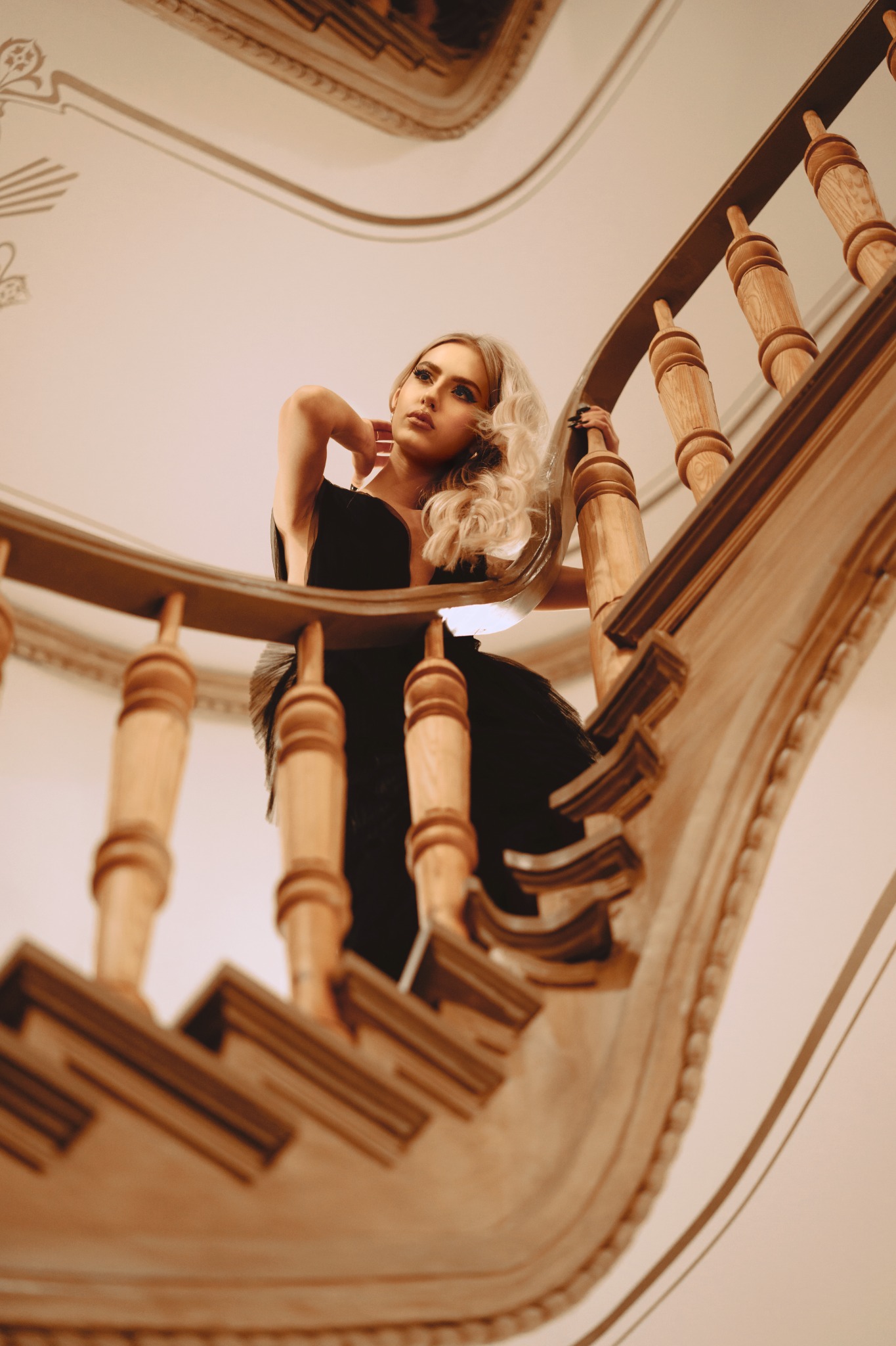 Blondwłosa kobieta w czarnej sukni pozuje na drewnianych kręconych schodach z dekoracyjnymi balustradami, patrząc w górę.