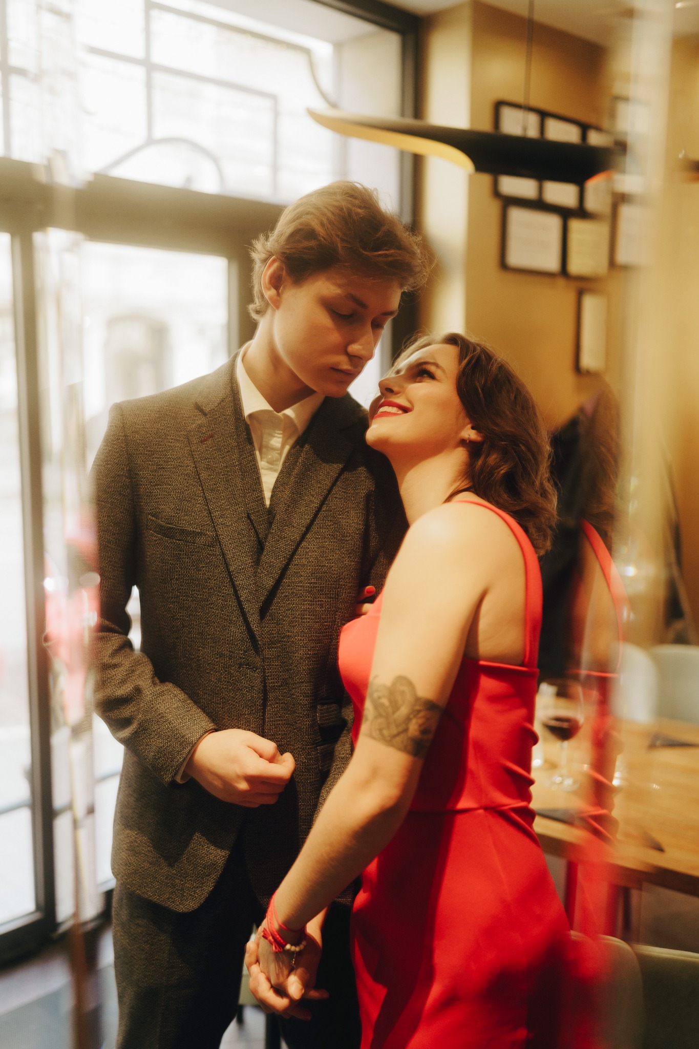 Zakochana para: mężczyzna w tweedowej marynarce i kobieta w czerwonej sukience, trzymają się za ręce w restauracji, ona patrzy na niego z uśmiechem, widoczny tatuaż na ramieniu kobiety.