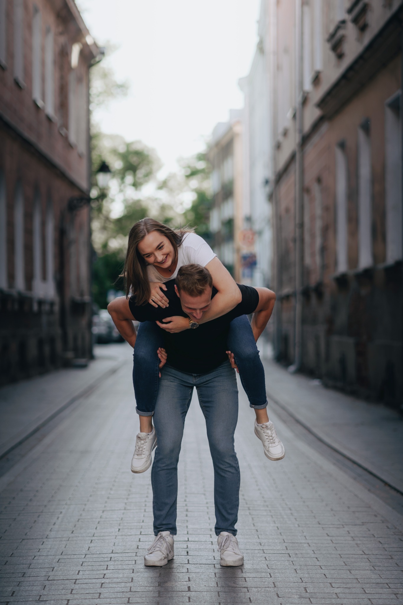 Uśmiechnięta kobieta na barana u mężczyzny na tle miejskiej uliczki z ceglanymi budynkami.