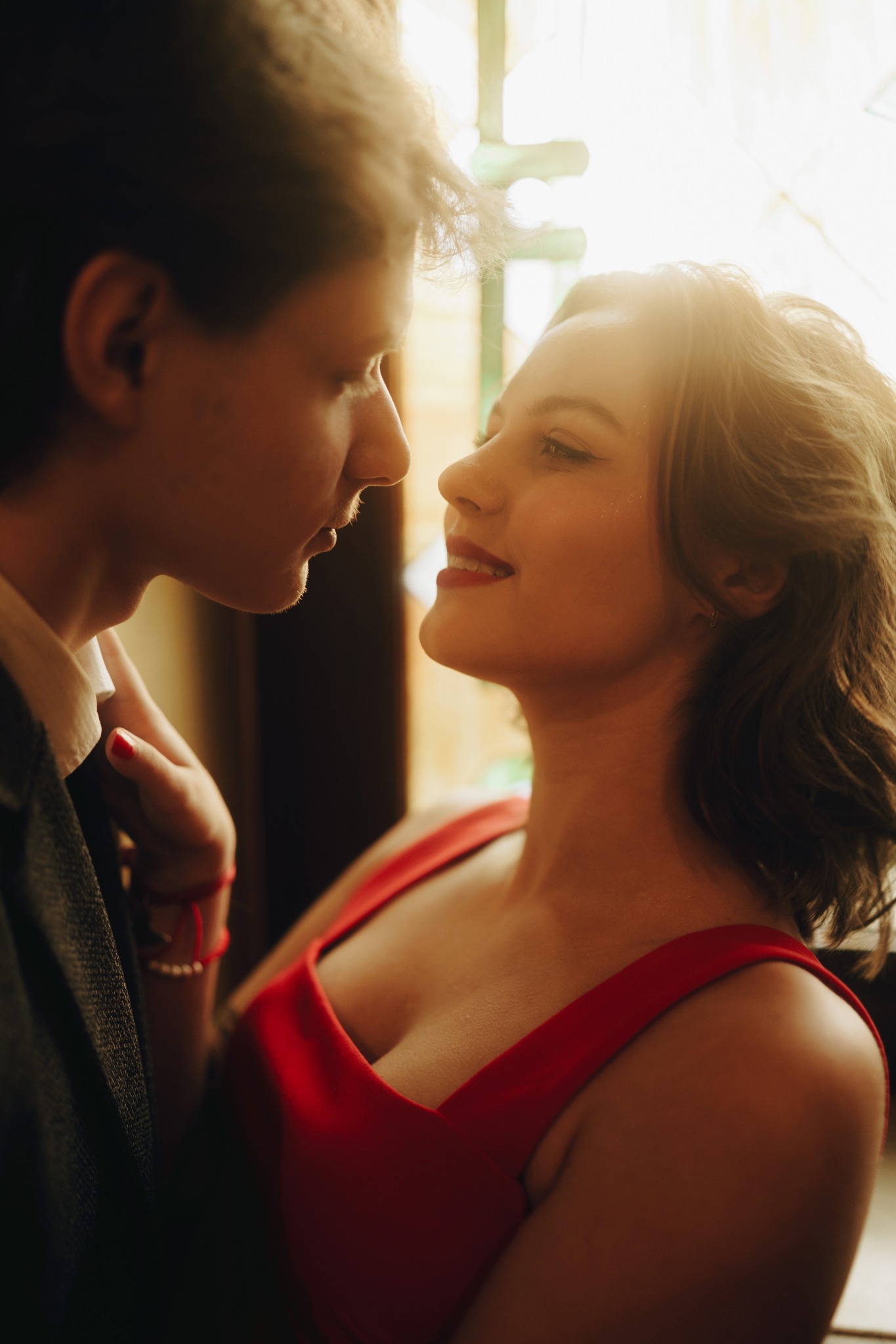 Para młoda w zbliżeniu, panna młoda w czerwonej sukience patrzy z uśmiechem na pana młodego, który trzyma ją za rękę, ciepłe oświetlenie.