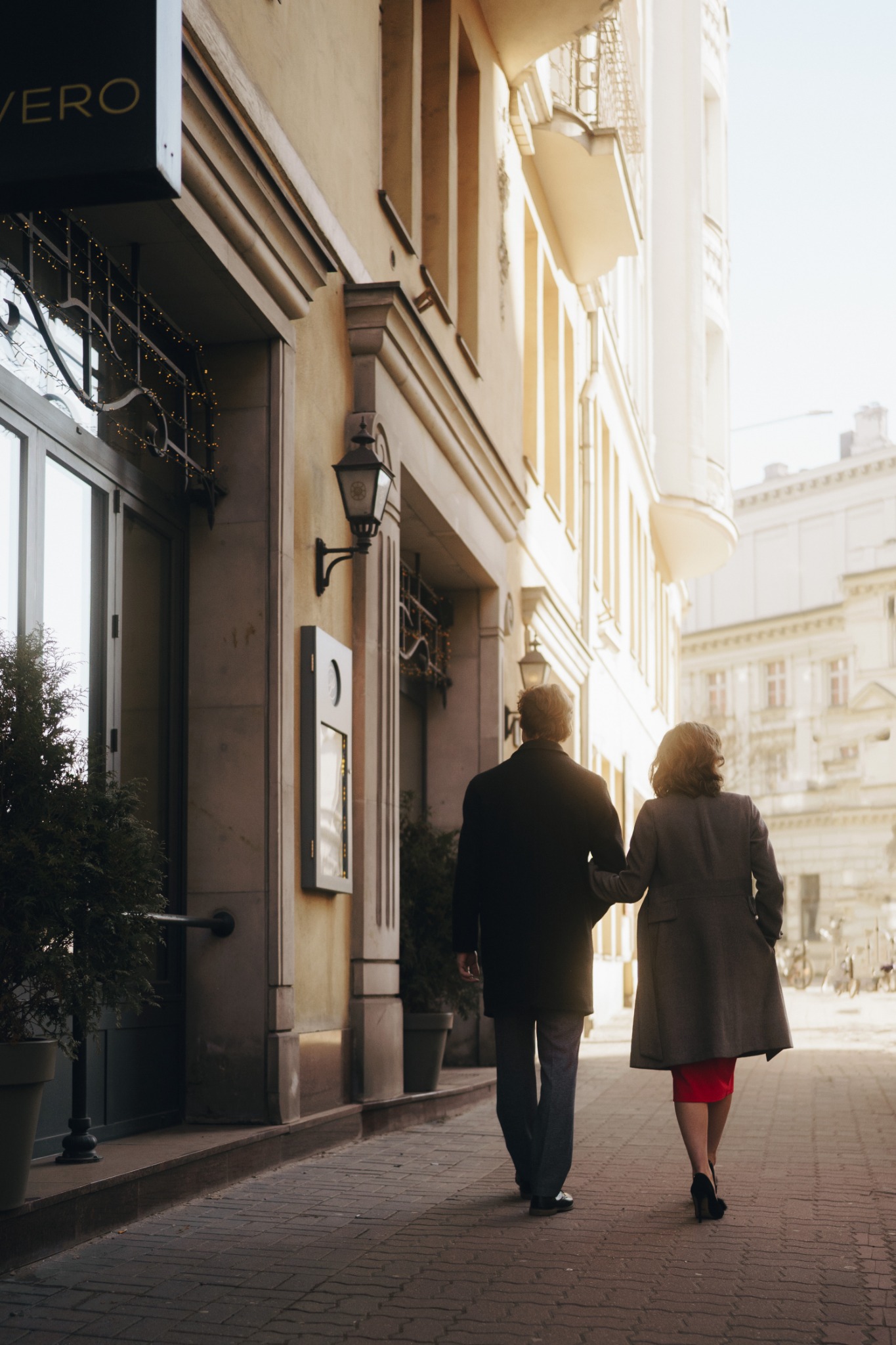 Para idzie ulicą obok budynku z szyldem 'VERO', trzymając się za ręce, kobieta w czerwonej sukience i szarym płaszczu, mężczyzna w ciemnym płaszczu i dżinsach.
