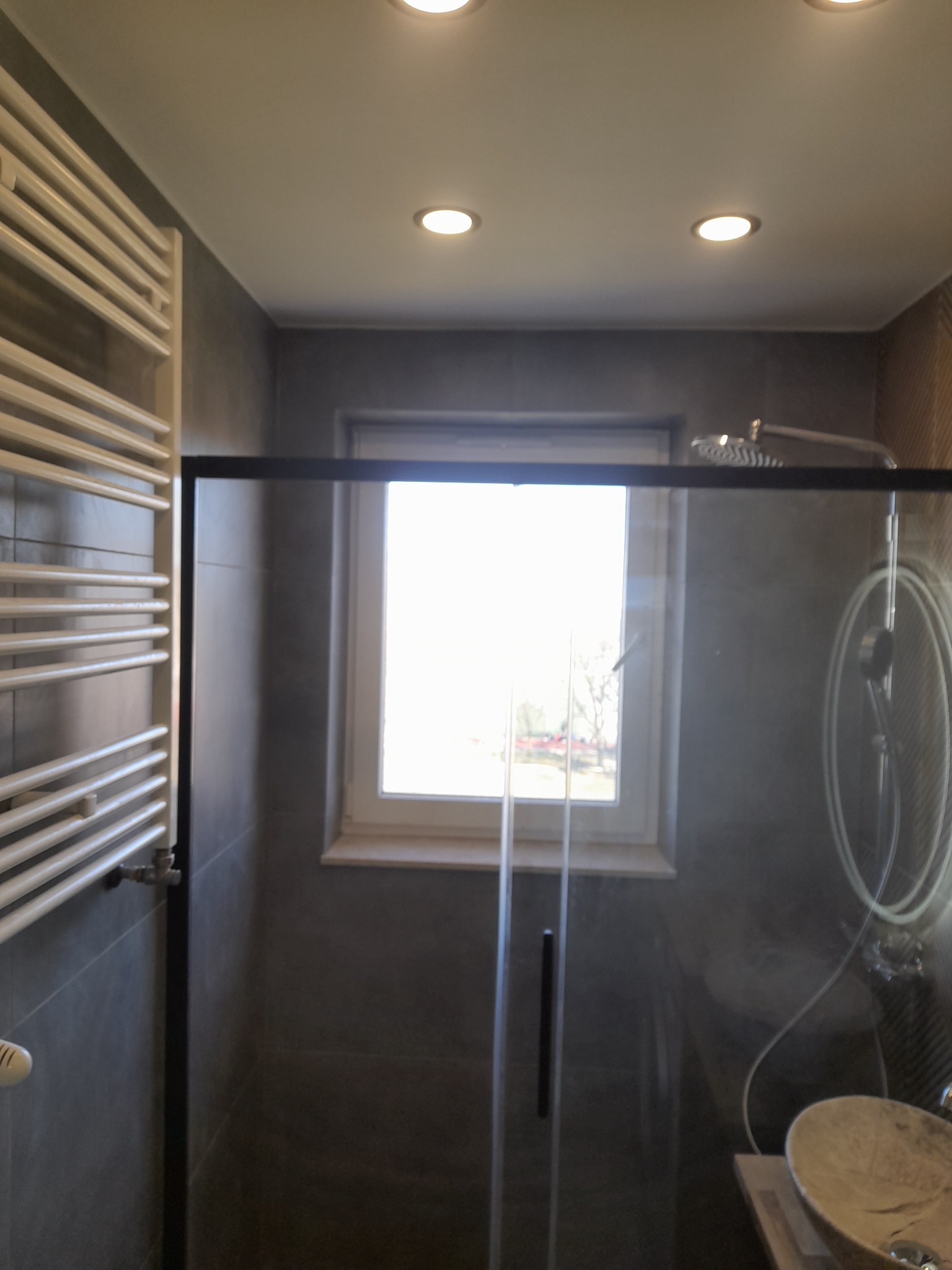 Nowoczesna łazienka z kabiną prysznicową typu walk-in, grzejnikiem drabinkowym i oknem. Szare płytki, punktowe oświetlenie sufitowe i umywalka nablatowa.