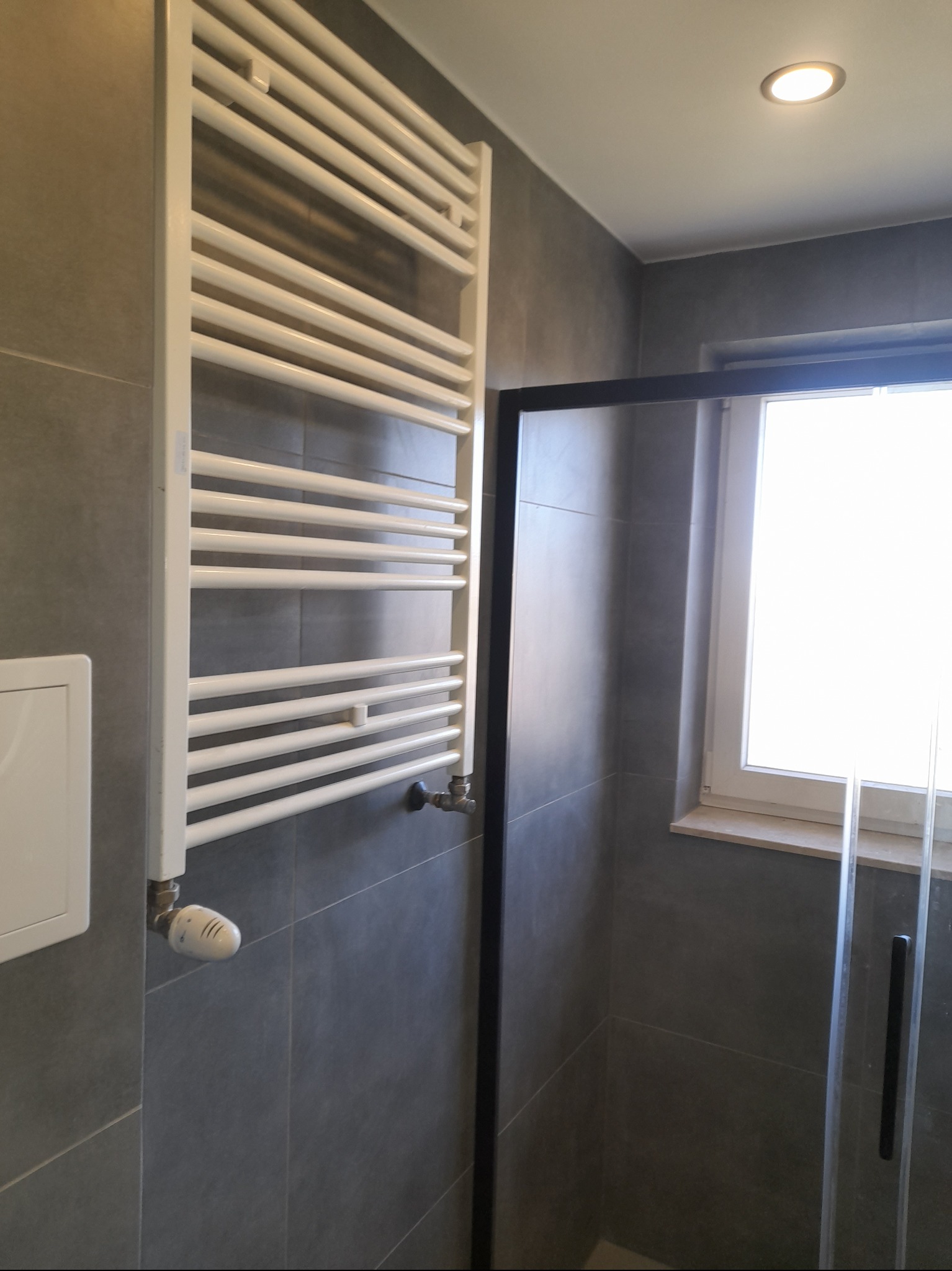 Szary, nowoczesny design łazienki z białym grzejnikiem drabinkowym i kabiną prysznicową z czarną ramą, oświetloną punktowym światłem.