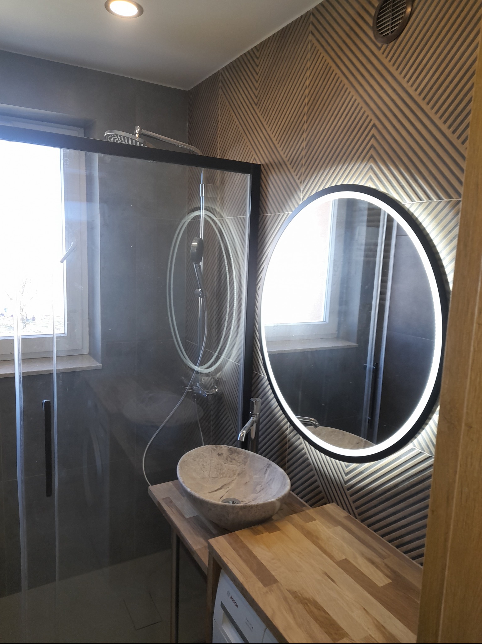 Nowoczesna łazienka z kabiną typu walk-in, umywalką z kamienia i okrągłym lustrem z podświetleniem LED. Geometryczny wzór na ścianie dodaje charakteru.