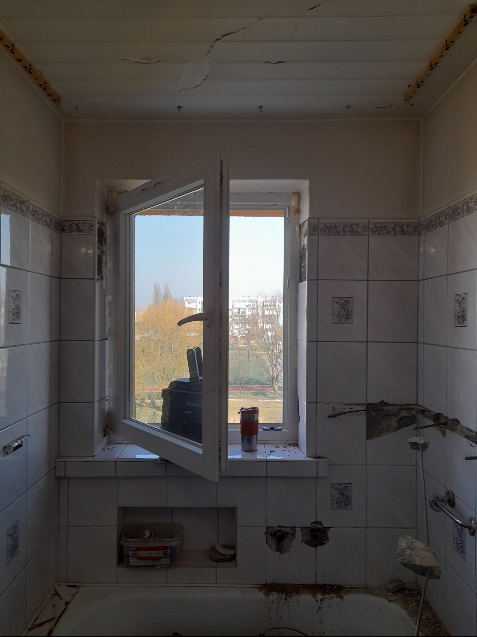 Łazienka w trakcie demontażu płytek, widoczne uszkodzenia na ścianach i suficie, otwarte okno z widokiem na zewnątrz, narzędzia i pojemniki na parapecie.
