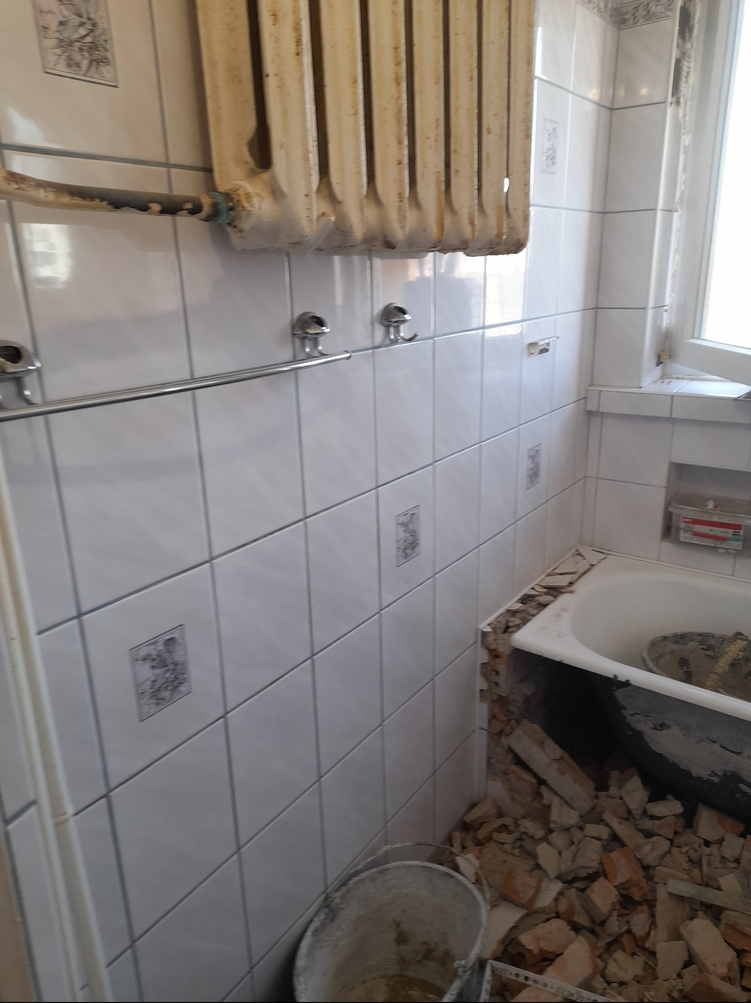 Demontaż starej łazienki: białe płytki z dekorami, żeliwny grzejnik, gruz wokół wanny. Widok na etap prac przygotowawczych do remontu.