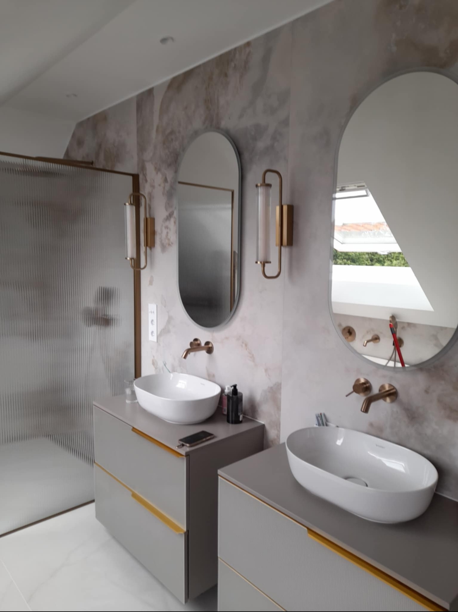 Stylowa łazienka z dwoma umywalkami nablatowymi, lustrami w owalnych ramach i złotymi akcentami. Ściany wykończone płytkami imitującymi marmur. Kabina prysznicowa z ryflowanego szkła.