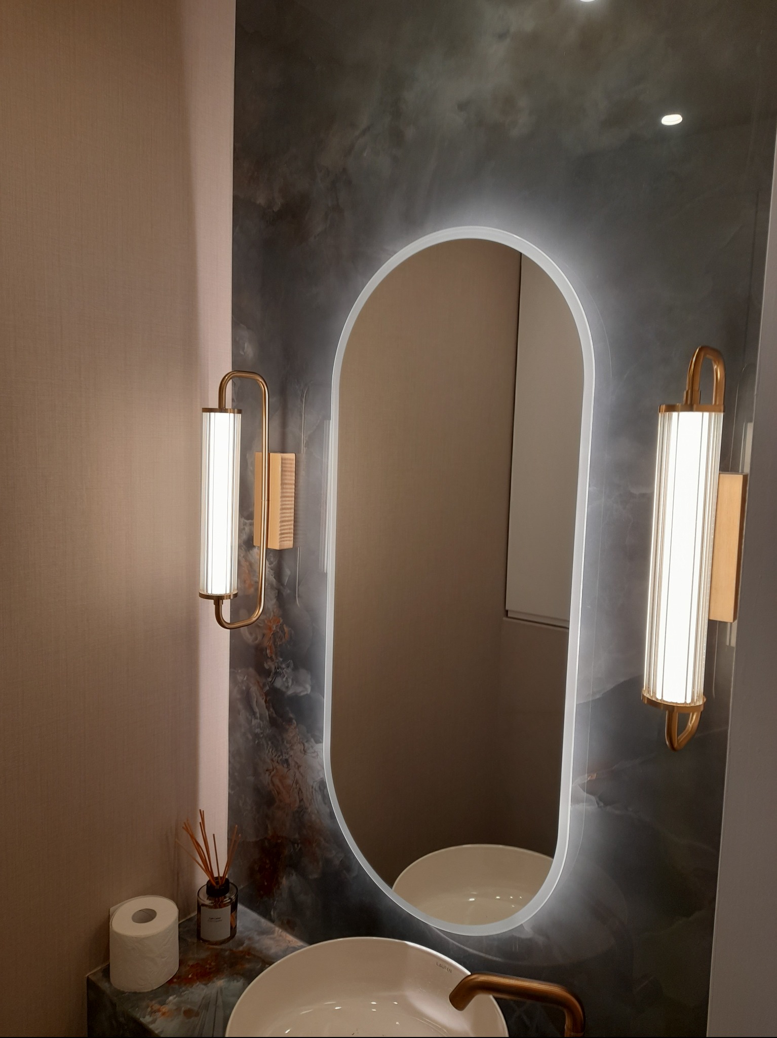 Nowoczesna łazienka z owalnym lustrem podświetlanym LED, umywalką nablatową i designerskimi lampami ściennymi. Marmurowa ściana i blat dodają elegancji.