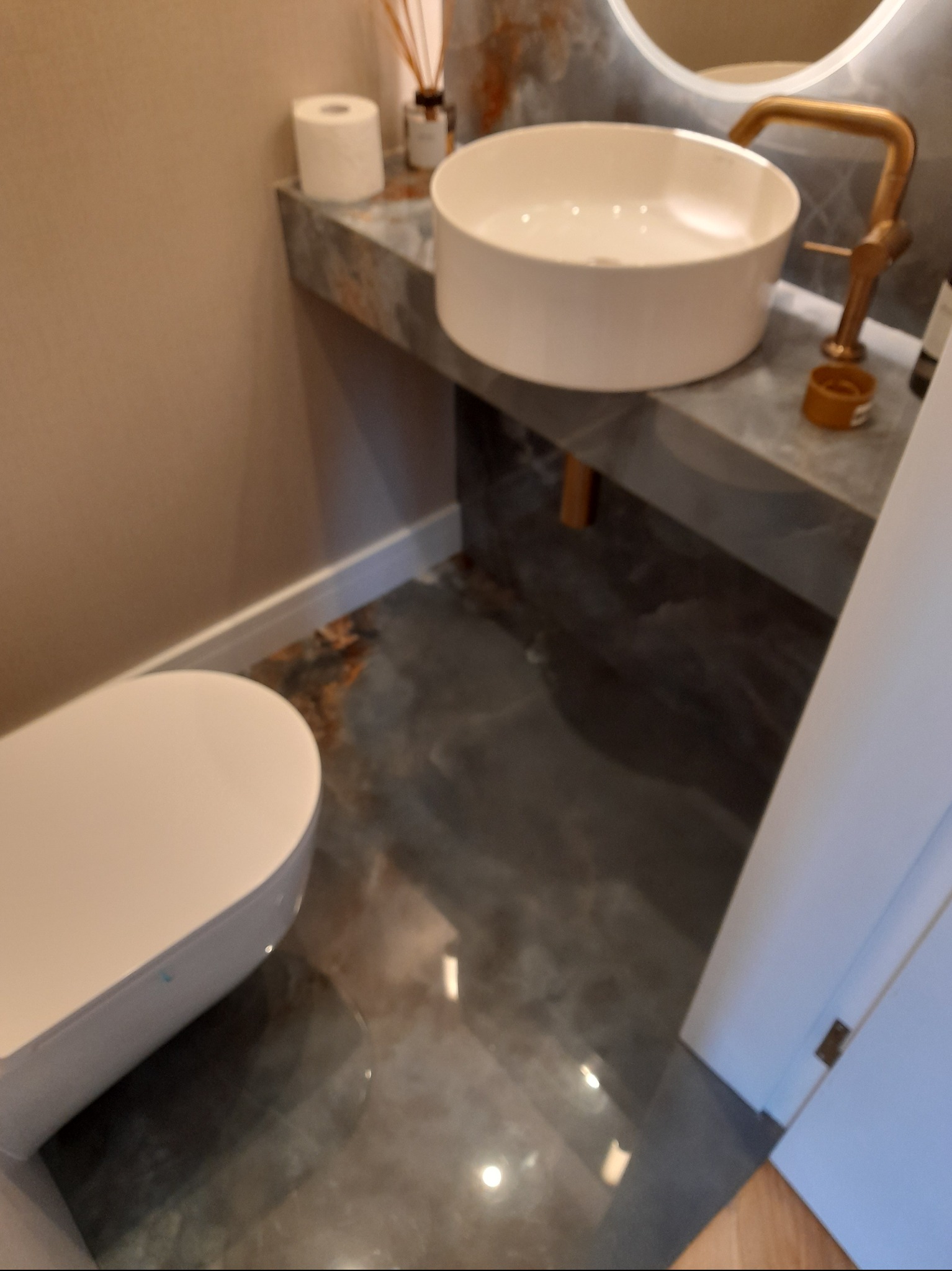 Nowoczesna łazienka z okrągłą umywalką nablatową, złotą armaturą i marmurowym blatem. Uwagę zwraca podłoga z efektem żywicy i minimalistyczna toaleta.