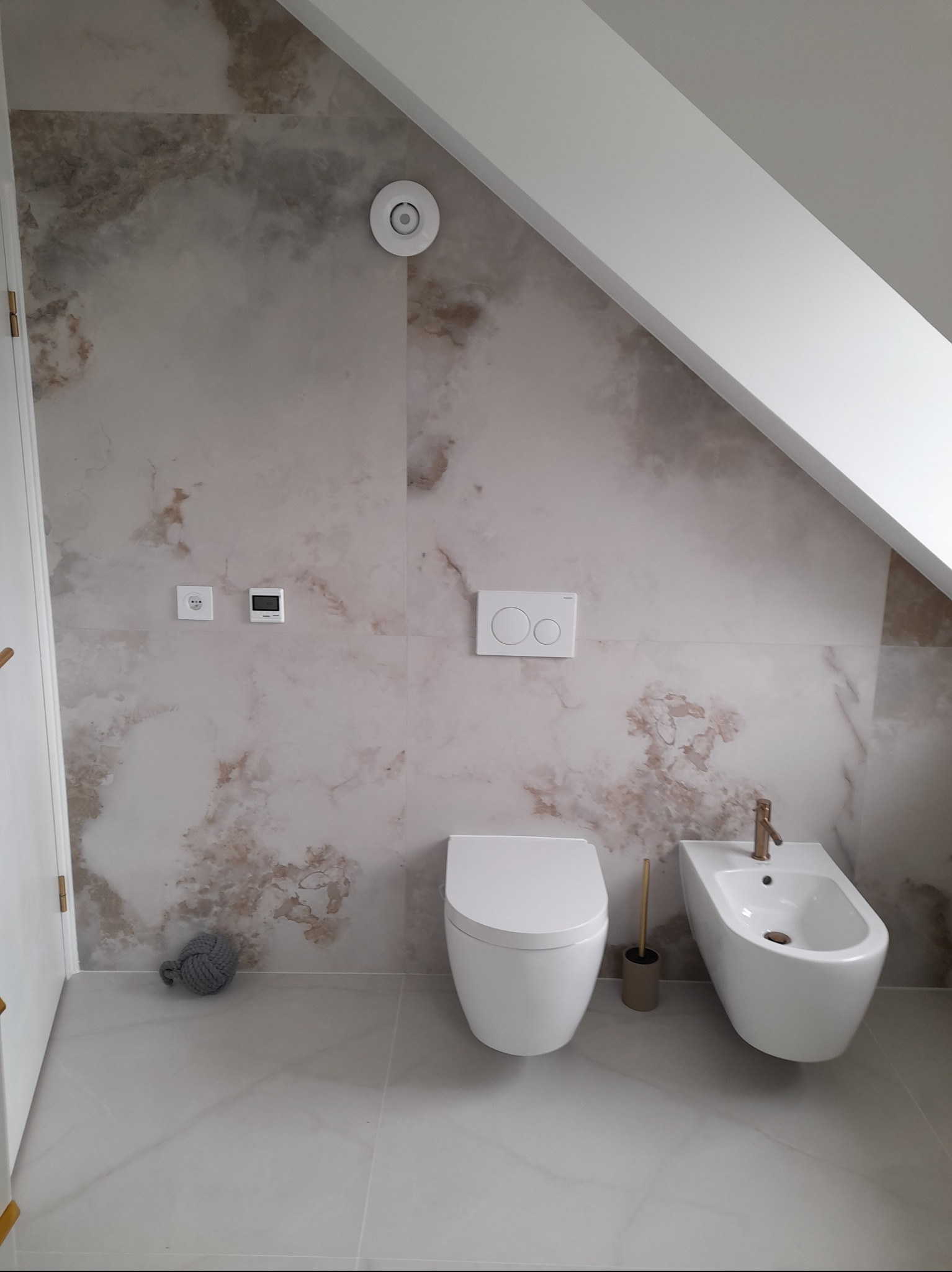 Wykończona łazienka na poddaszu: marmurowe płytki, biała toaleta i bidet z miedzianą armaturą. Minimalistyczny design, widoczne skosy.