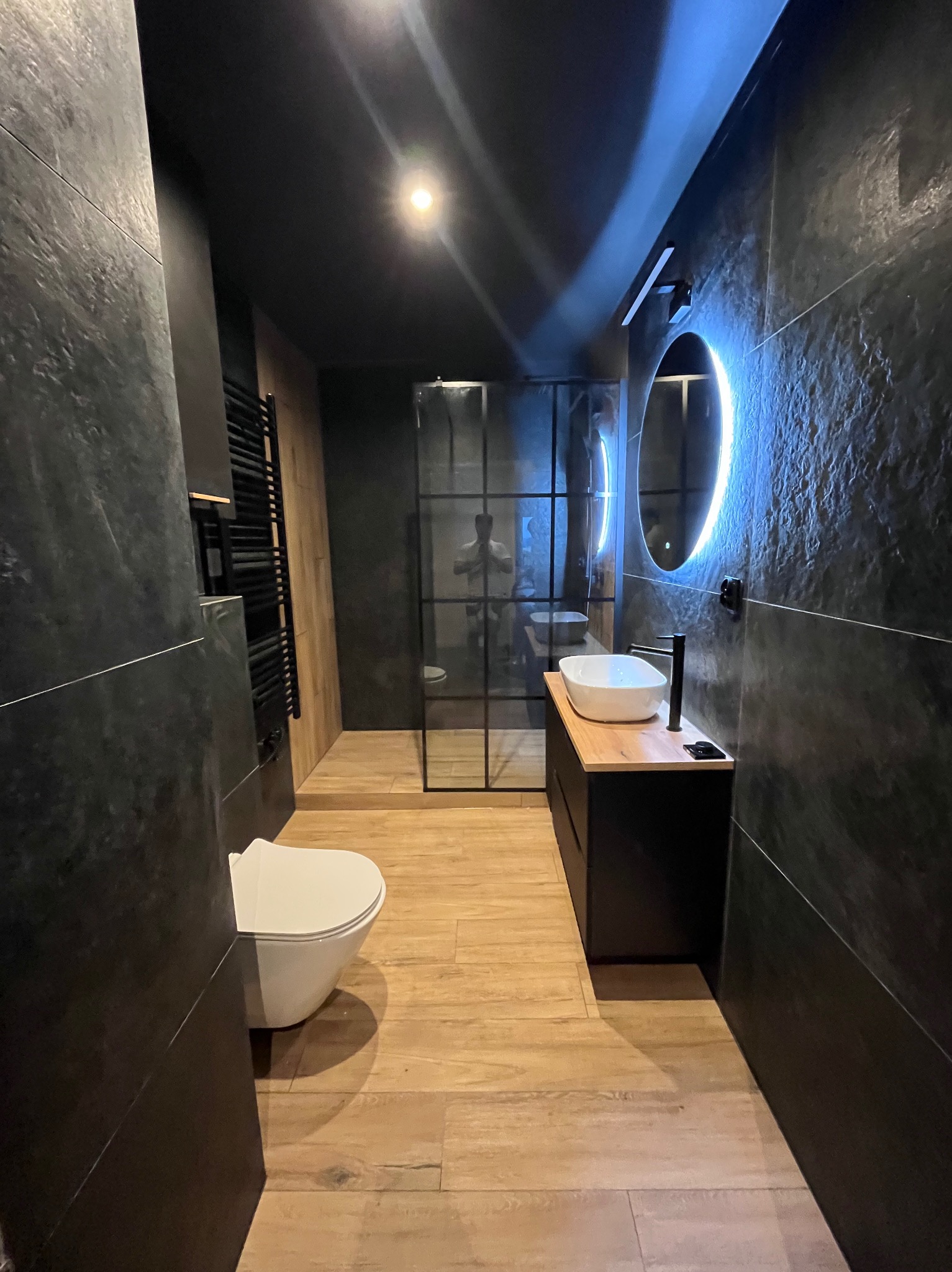 Nowoczesna łazienka w ciemnych tonacjach z drewnianą podłogą, kabiną prysznicową typu walk-in i okrągłym lustrem z podświetleniem LED. Minimalistyczny design.