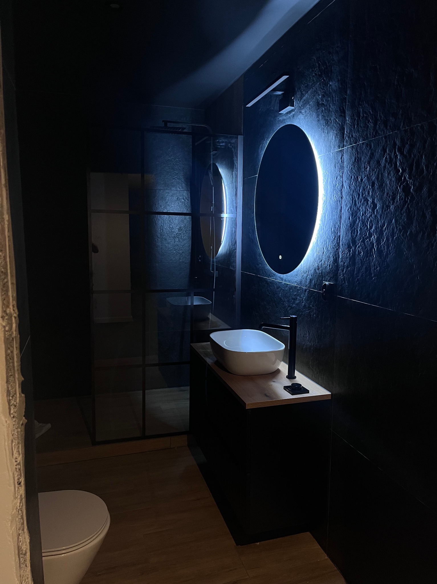 Nowoczesna łazienka z czarnymi płytkami o nieregularnej fakturze, podświetlanym okrągłym lustrem i kabiną prysznicową ze szklanymi ściankami.