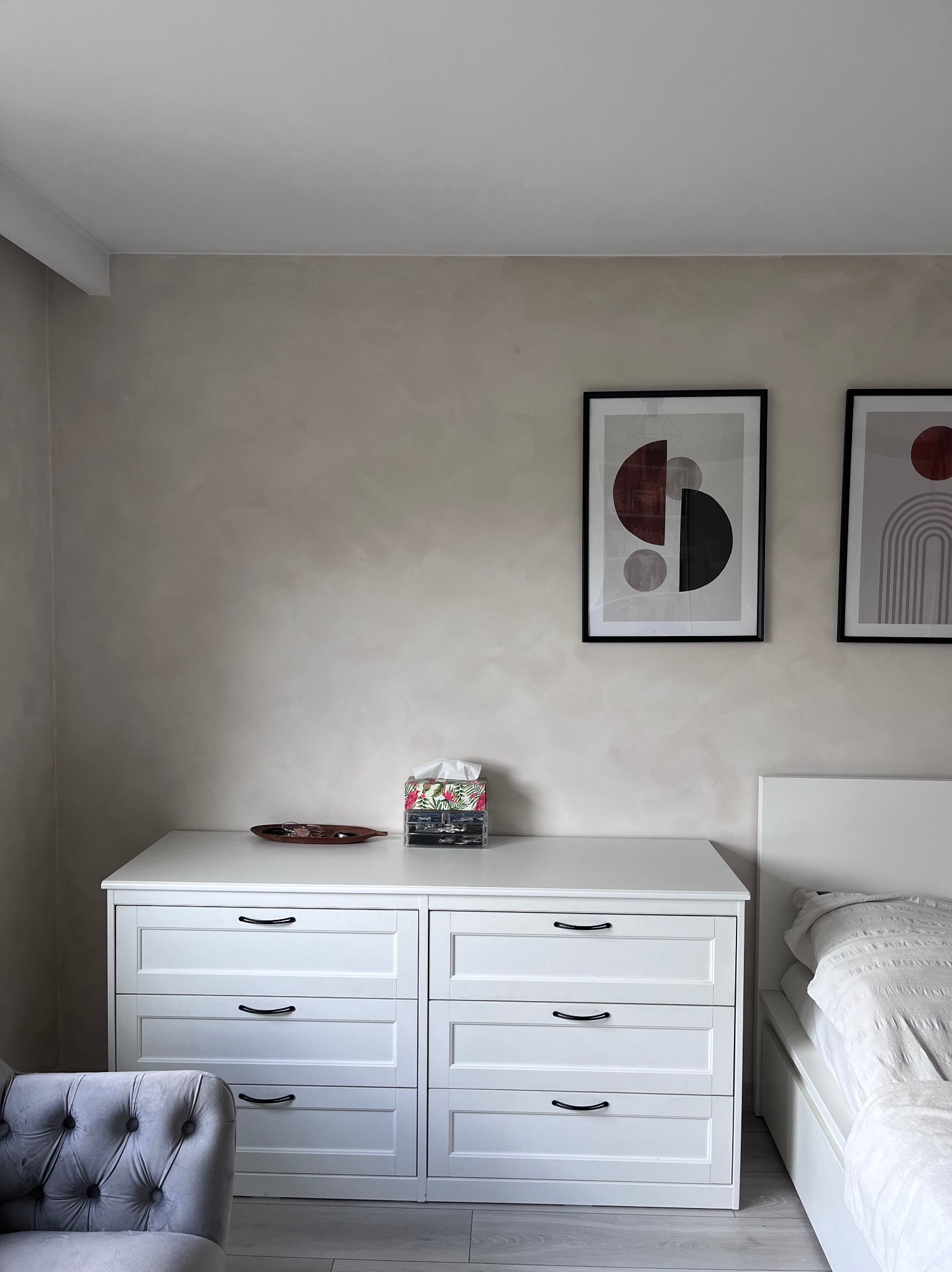 Ściana z efektem dekoracyjnym w beżowym odcieniu, nad białą komodą z czarnymi uchwytami. Na ścianie wiszą dwa obrazy w czarnych ramkach z motywami geometrycznymi.