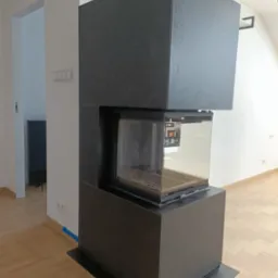 Nowoczesny, narożny kominek z czarną obudową w surowym wnętrzu, widoczne detale wykończenia i odblask w szybie.