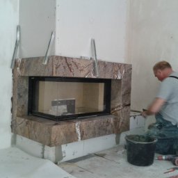 Kominki Mikam - Montaż obudowy kominka z brązowego kamienia z wyraźnym wzorem żyłkowania, widoczne metalowe profile montażowe, pracownik w kombinezonie wygładza zaprawę, wiadro z zaprawą i narzędzia na podłodze.