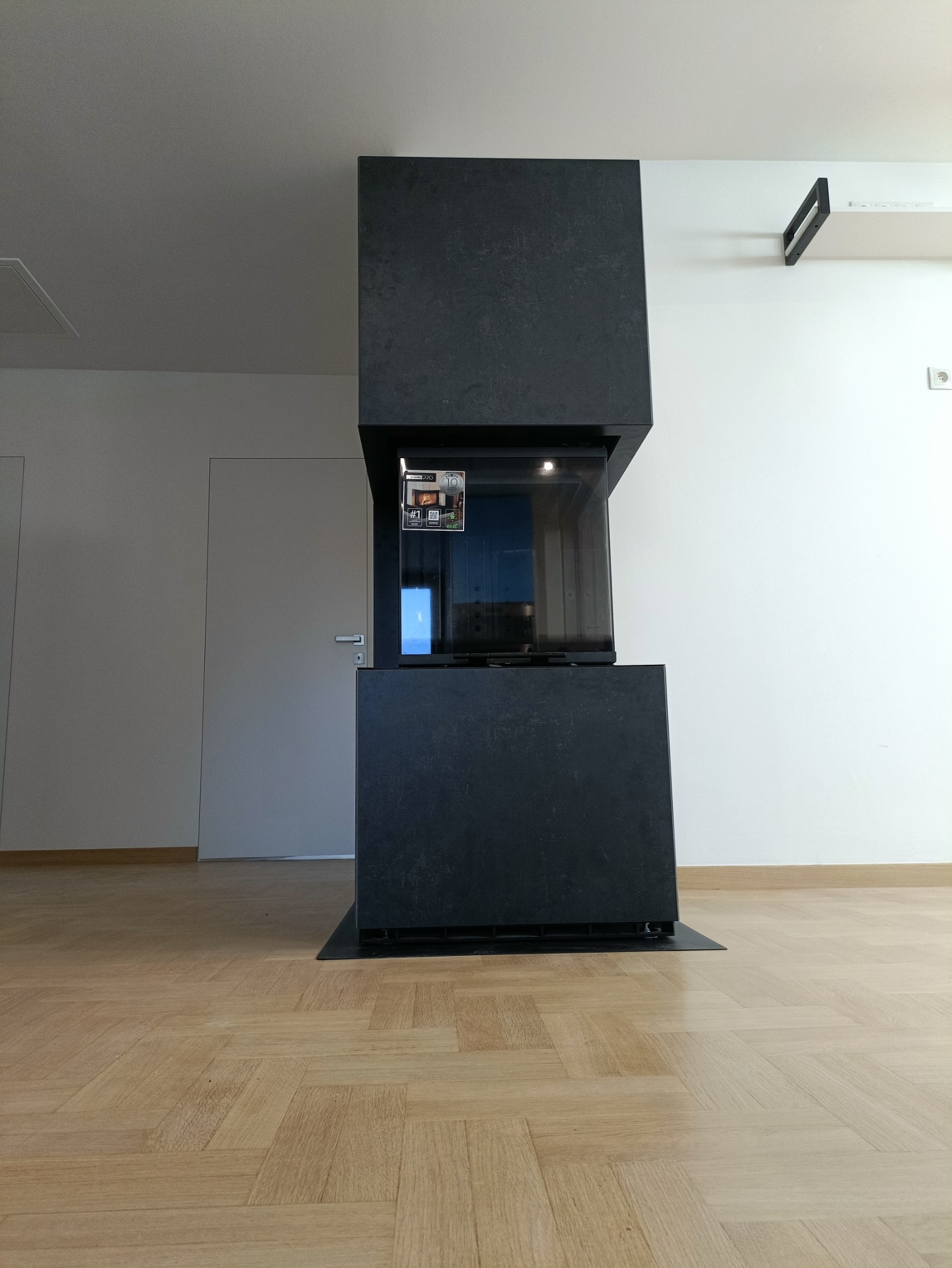 Nowoczesny, czarny kominek z przeszklonym paleniskiem, zamontowany w jasnym wnętrzu w Katowicach, widoczny na tle białej ściany i parkietu ułożonego w jodełkę.