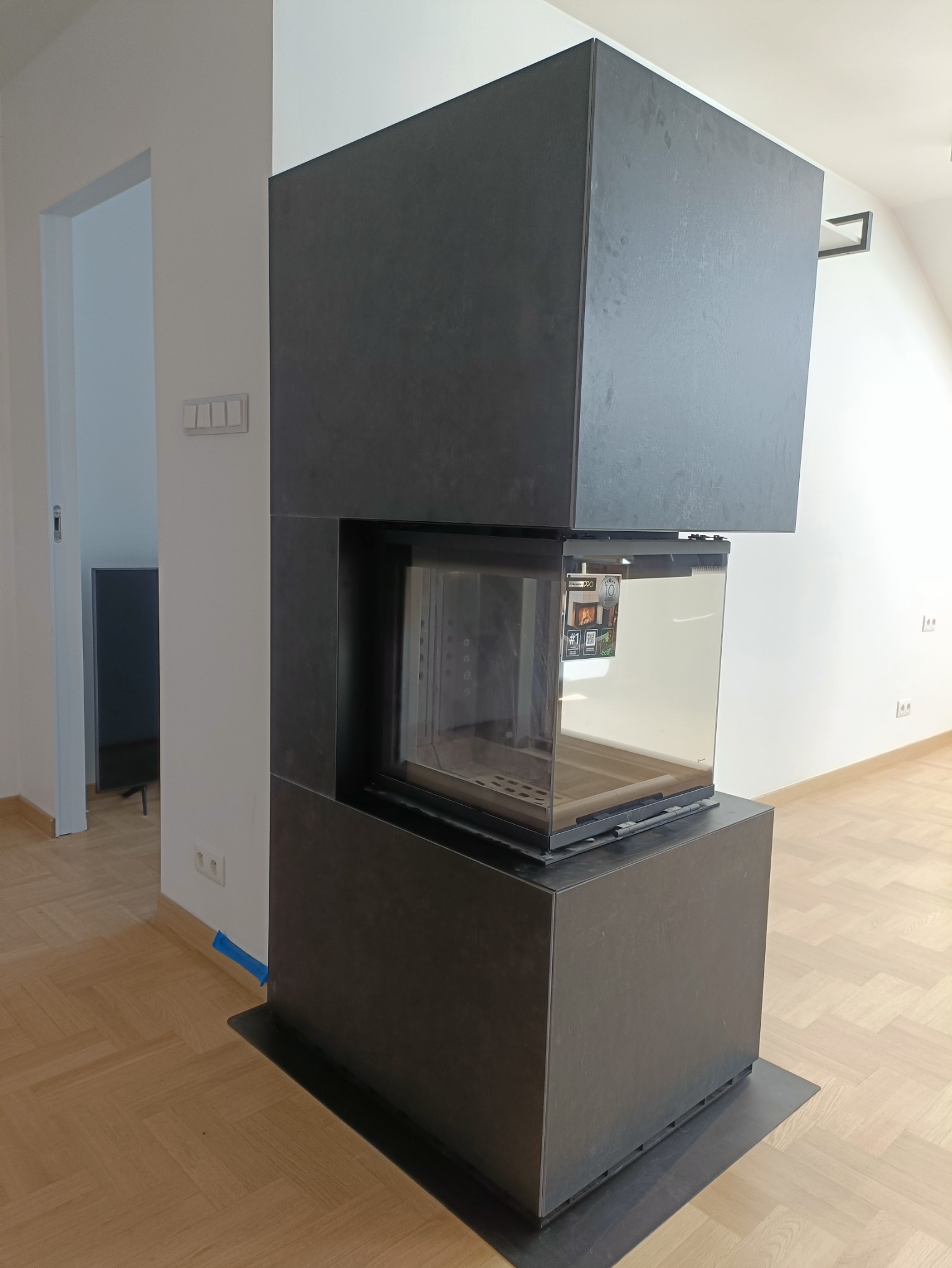 Nowoczesny, narożny kominek z czarną obudową w surowym wnętrzu, widoczne detale wykończenia i odblask w szybie.