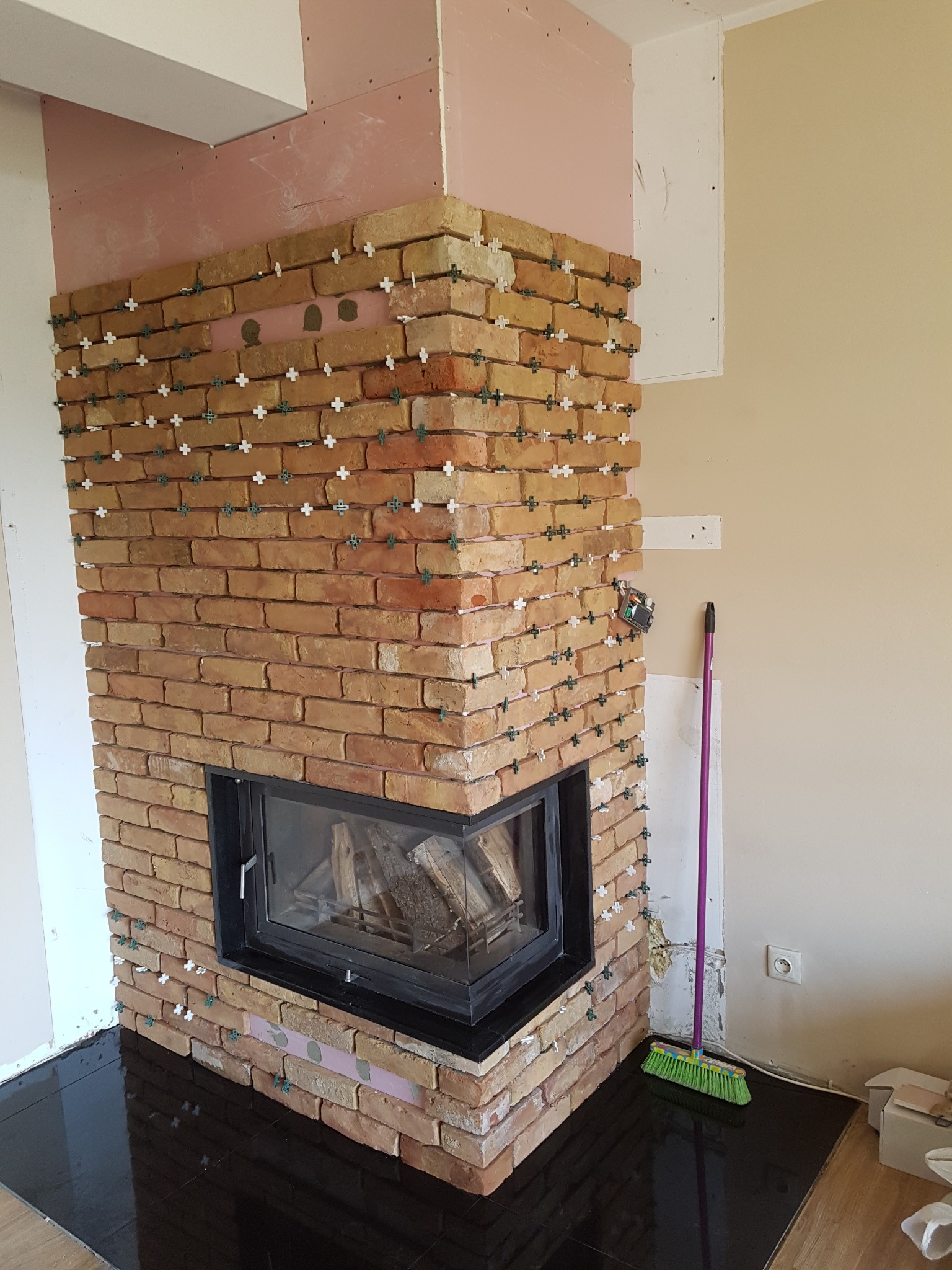 Murowany kominek z cegły klinkierowej w trakcie budowy, widoczne krzyżyki dystansowe między cegłami, czarna wkładka kominkowa oraz przygotowane ściany do dalszych prac wykończeniowych.