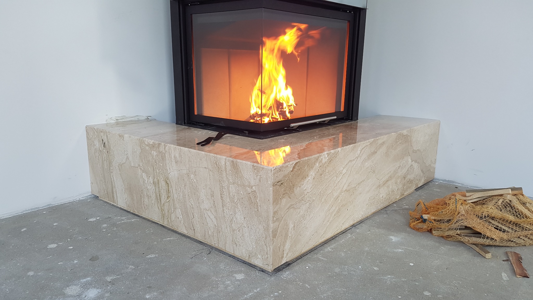 Nowoczesny kominek narożny z obudową z trawertynu, płomienie widoczne za szybą, obok siatka z drewnem na surowym betonie.