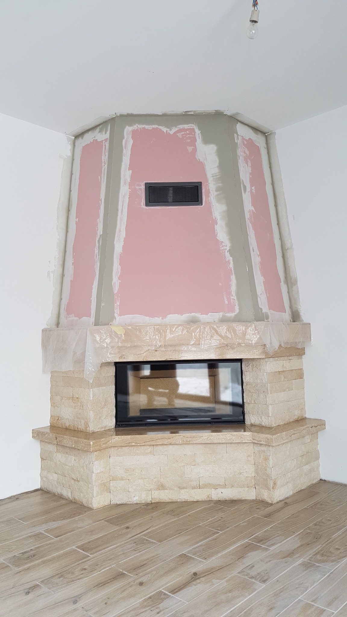 Kominek w trakcie wykończenia: obudowa z kamienia naturalnego, różowe płyty karton-gips na górze, zabezpieczone folią, surowy stan ścian i podłogi.