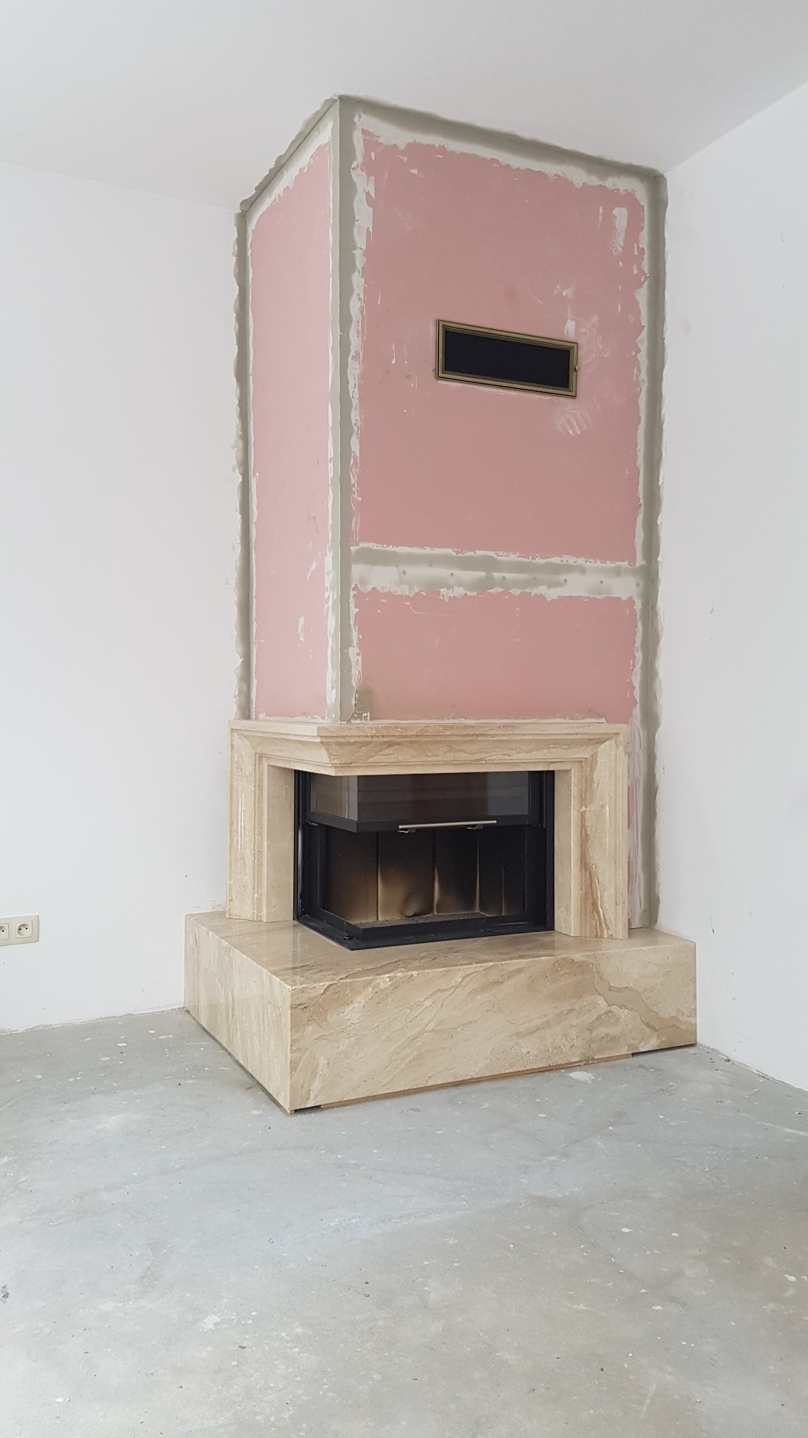 Wnętrze z kominkiem w trakcie zabudowy: obudowa z marmuru i ściany z płyt kartonowo-gipsowych przed wykończeniem, widoczna kratka wentylacyjna.