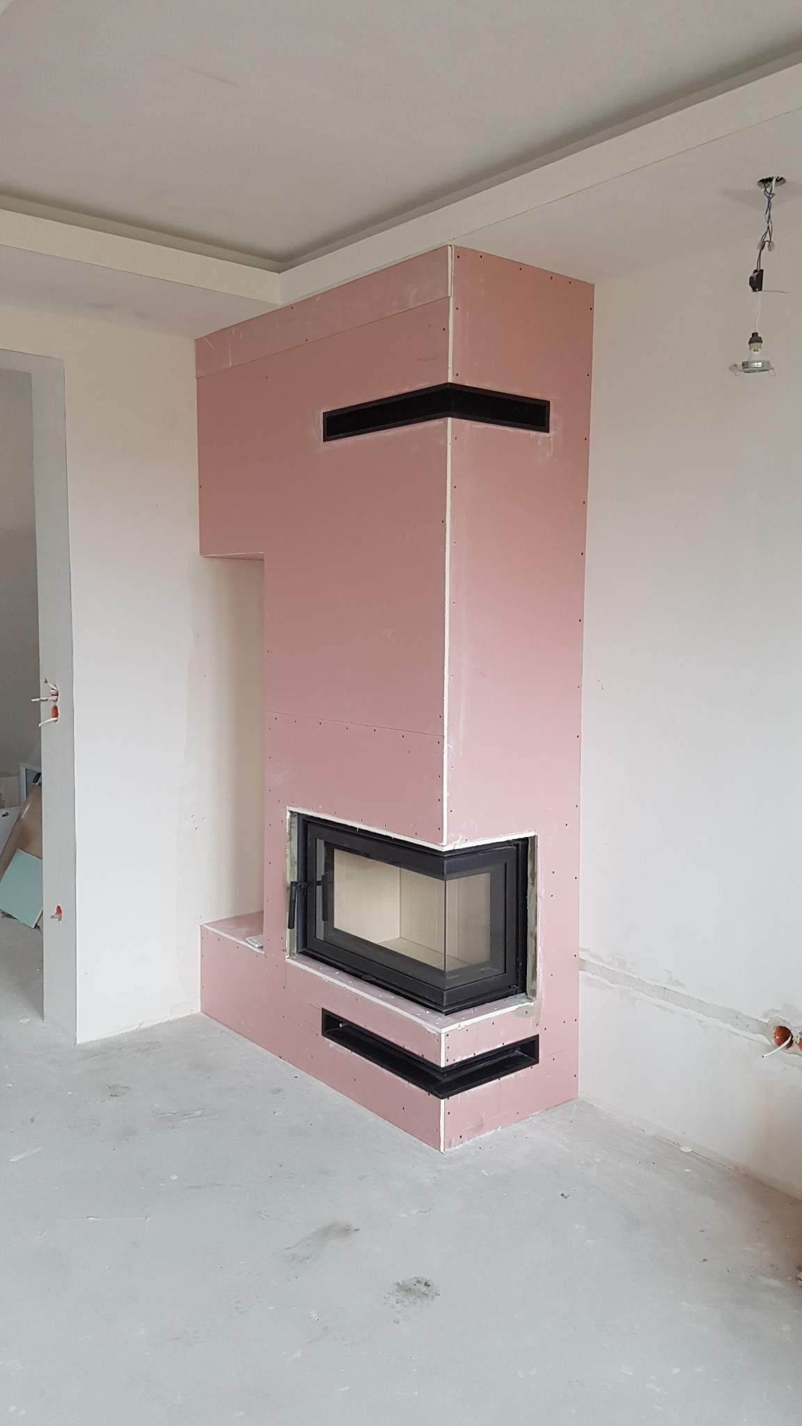 Niedokończona obudowa kominka z płyty kartonowo-gipsowej, z widocznymi otworami wentylacyjnymi, czarnym wkładem kominkowym, surowym betonem na podłodze i wiszącą lampą bez klosza.