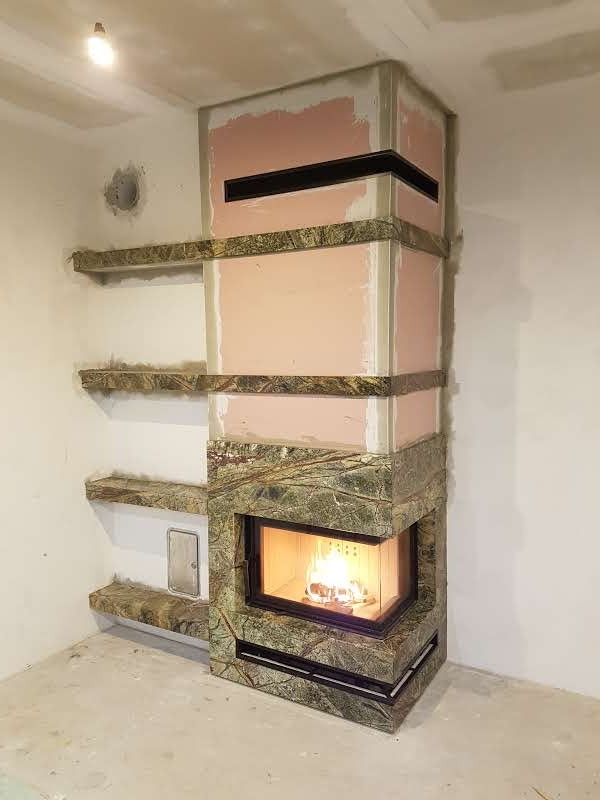 Niedokończona obudowa kominka z zielonego marmuru z płomieniami widocznymi przez szybę, z półkami w tym samym stylu i surowym wykończeniem ścian.