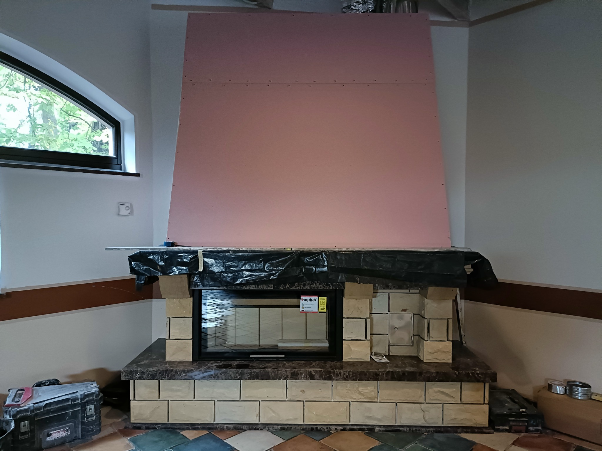 Budowa kominka: widok na wkład kominkowy obudowany cegłami, zabezpieczony folią, z zamontowaną płytą kartonowo-gipsową na górze, narzędzia w skrzynce obok.