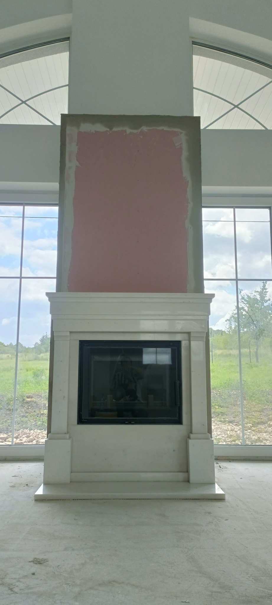 Wnętrze w Katowicach z niedokończonym kominkiem z białym obramowaniem, ściana nad nim przygotowana pod malowanie, duże okna z widokiem na zewnątrz.