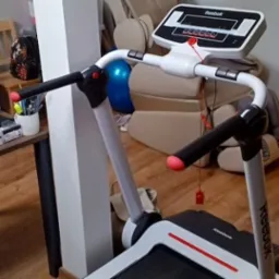 Bieżnia marki Reebok z czarnymi uchwytami i czerwonymi akcentami, w tle niebieska piłka do ćwiczeń i beżowy fotel masujący.