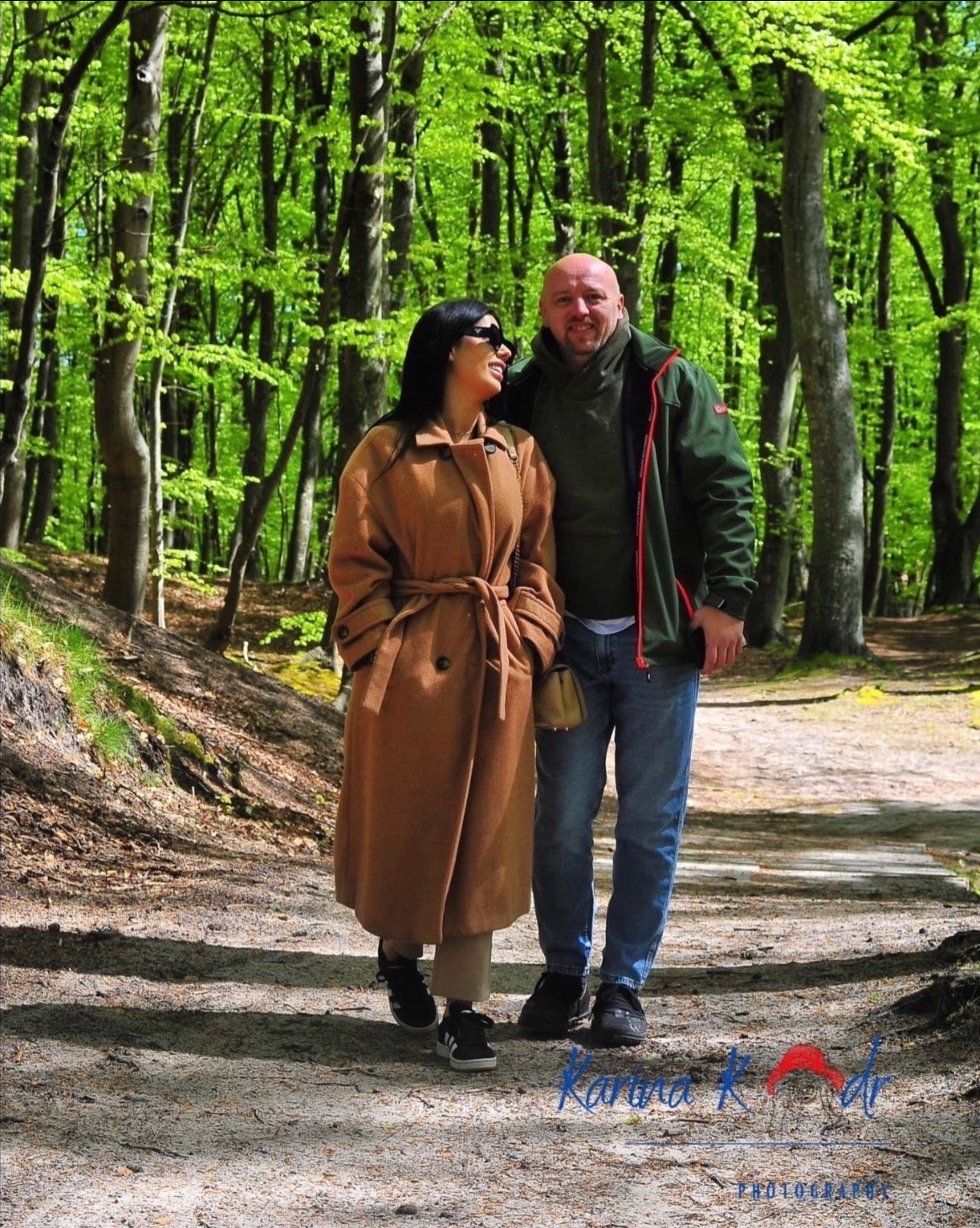 Para w jesiennych strojach spaceruje po leśnej ścieżce w słoneczny dzień, drzewa w tle mają intensywnie zielone liście.