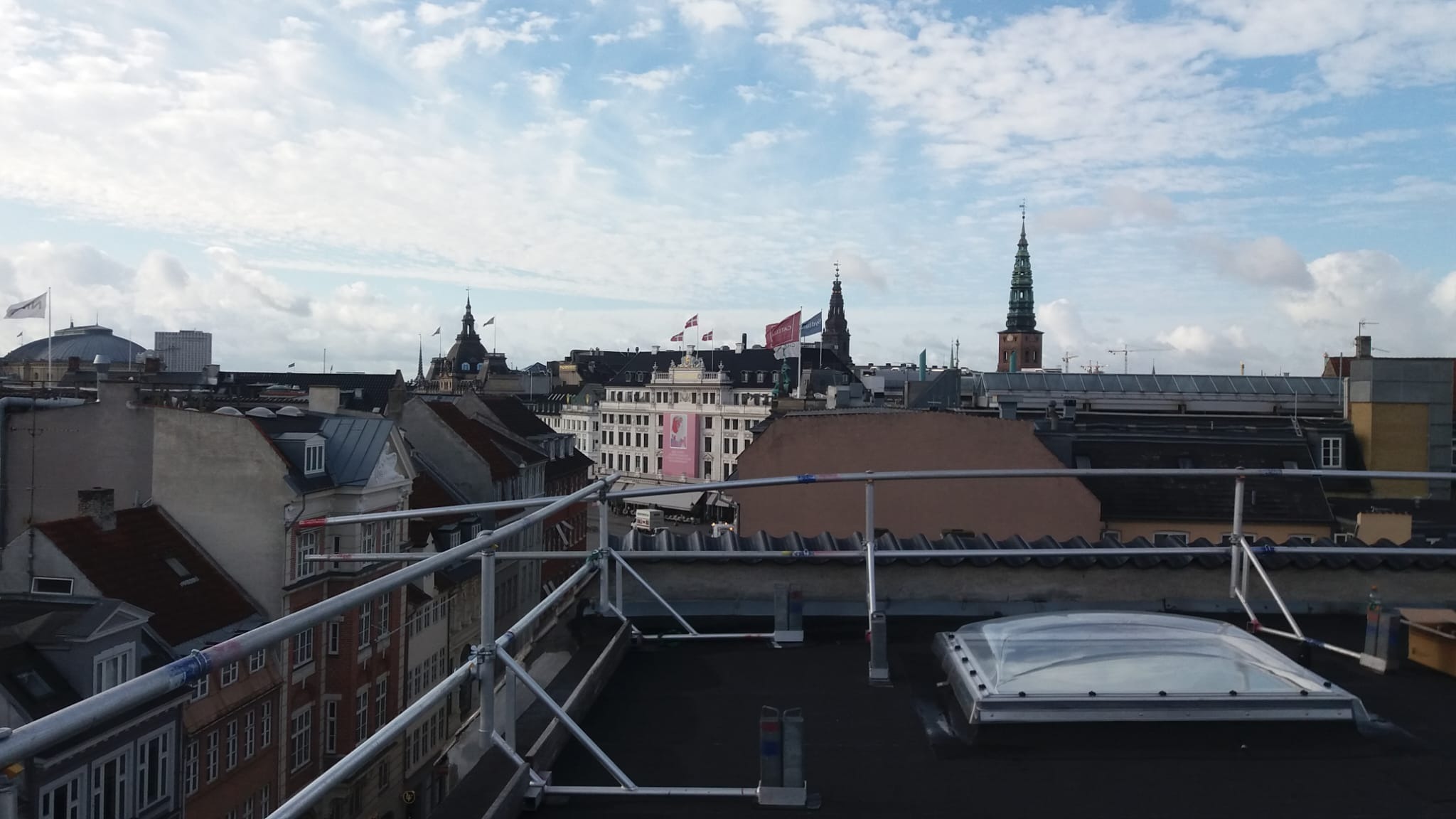 Widok z dachu na panoramę miasta z rusztowaniem i świetlikiem dachowym, widoczne budynki z flagami i wieżami na tle pochmurnego nieba.