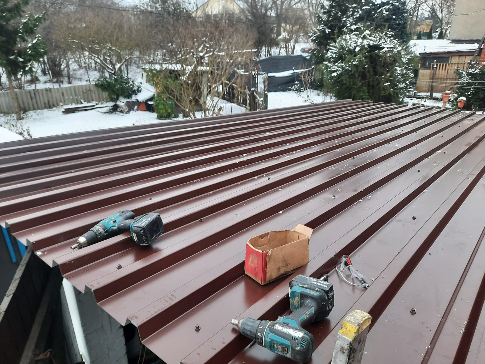Brązowy dach z blachy trapezowej w trakcie montażu, widoczne wkrętarki akumulatorowe, kartonowe pudełko i narzędzia, w tle ośnieżone drzewa i budynki.