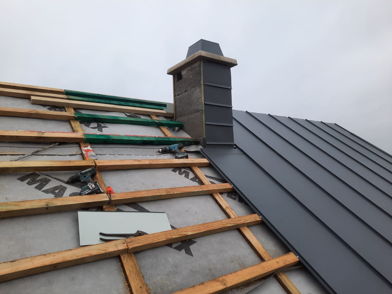 Montaż blachy na dachu w trakcie realizacji, widoczne drewniane łaty, membrana dachowa, narzędzia oraz fragment komina