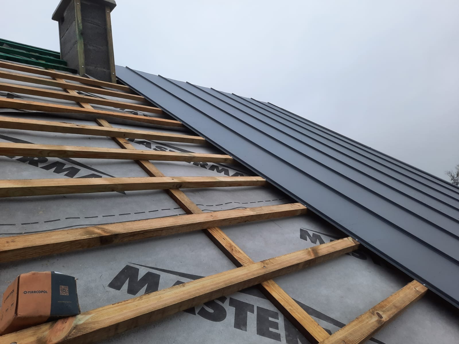 Montaż blachy na dachu: widoczne drewniane krokwie, membrana dachowa z nadrukiem i częściowo ułożone panele blaszane w kolorze grafitowym, komin w tle.
