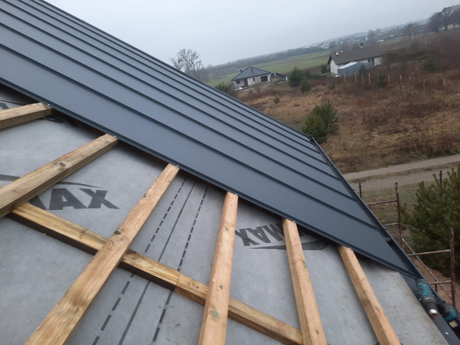Montaż grafitowej blachy na dachu z widoczną konstrukcją drewnianą i membraną, w tle krajobraz z domami.