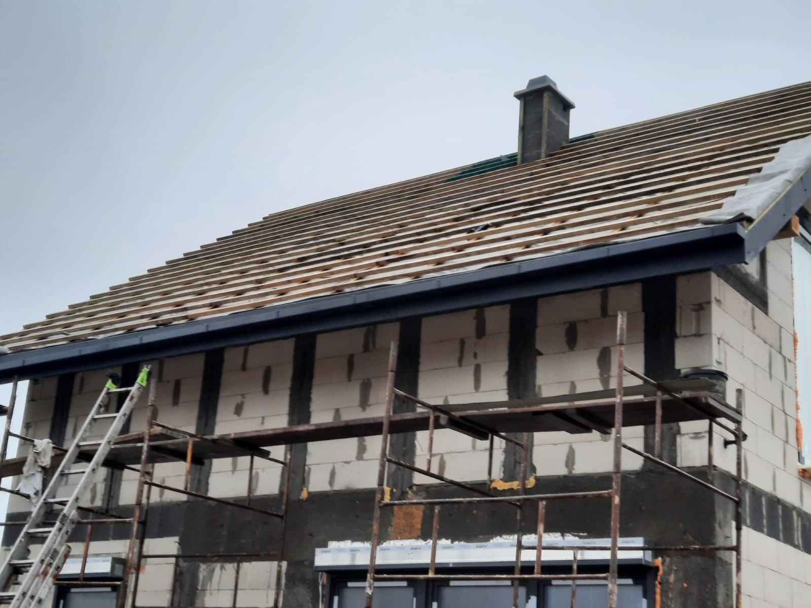 Dom w trakcie budowy z widocznym przygotowaniem dachu: ułożone łaty pod pokrycie, zamontowane rynny w kolorze grafitowym, fragment komina. Widoczne rusztowanie i drabina.