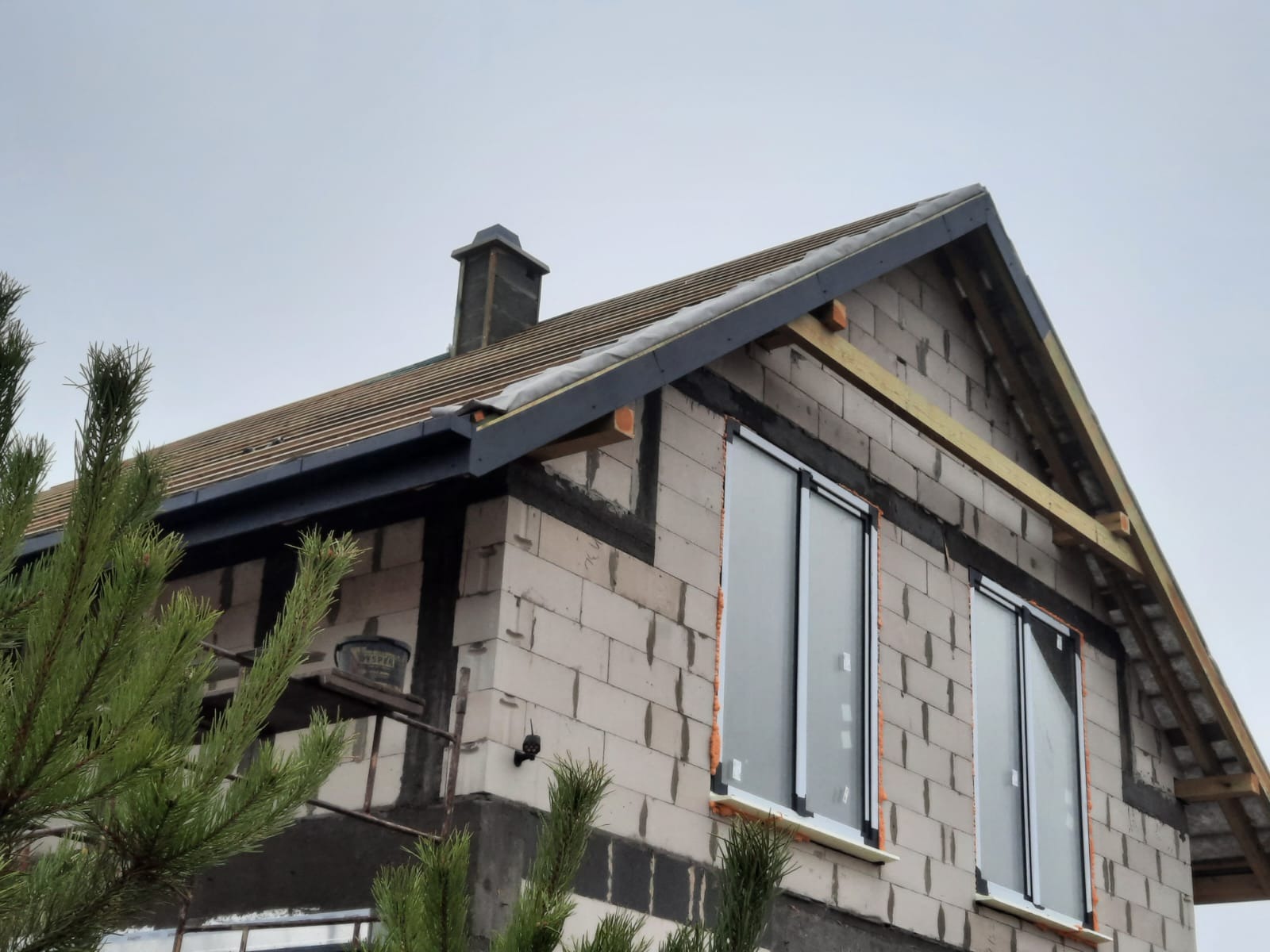 Dom w trakcie budowy z widocznym dachem pokrytym dachówką, kominem i oknami z pianką montażową, na tle pochmurnego nieba.