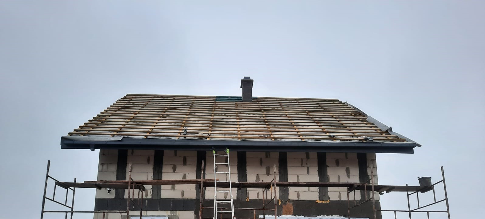 Budynek w trakcie krycia dachu, widoczne łaty pod dachówkę, komin i rusztowania.