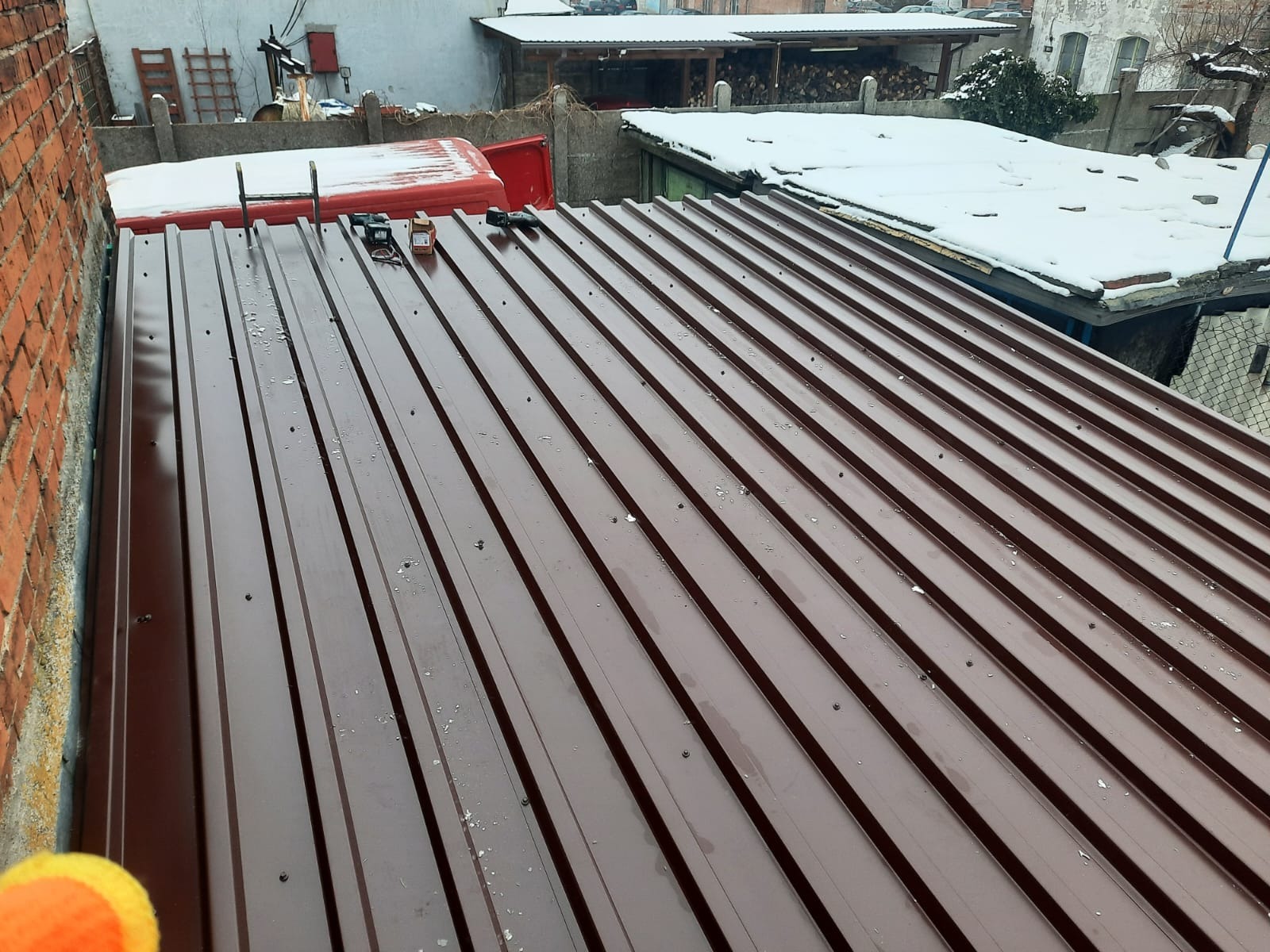 Nowe, brązowe pokrycie dachowe z blachy trapezowej, widoczne narzędzia dekarskie i fragment czerwonego samochodu na tle zabudowań w zimowej scenerii.