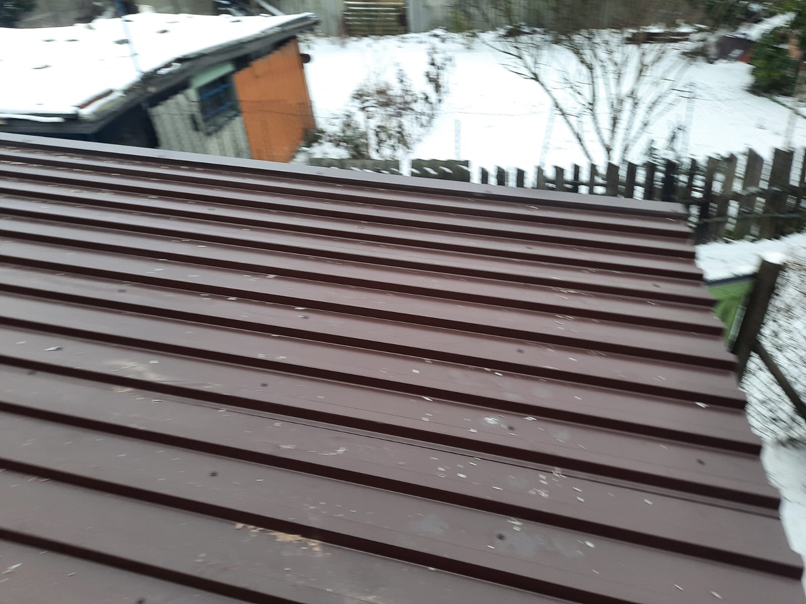 Nowe, brązowe pokrycie dachowe z blachy trapezowej na tle ośnieżonego ogrodu i zabudowań gospodarczych.