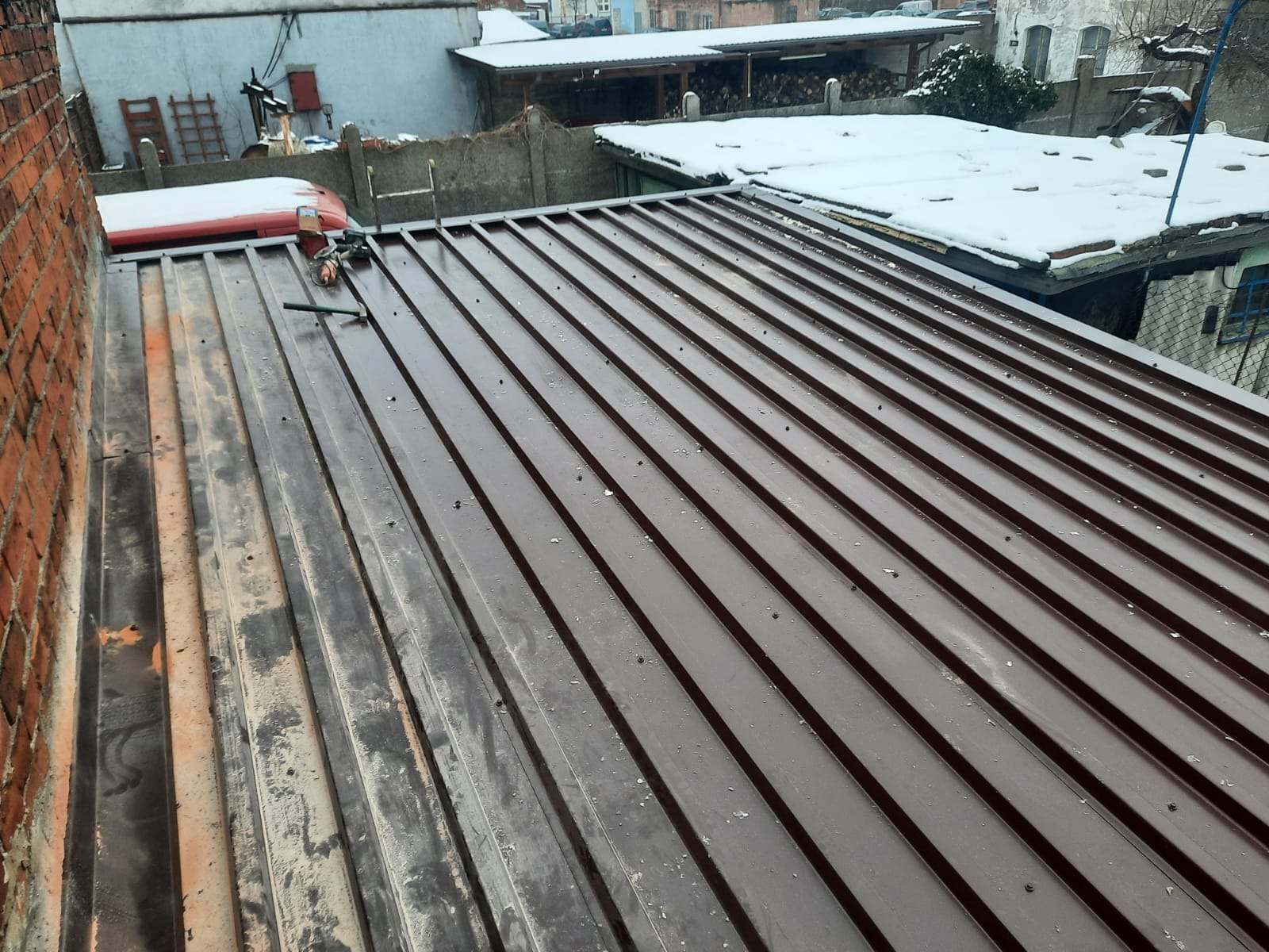 Nowy, brązowy dach z blachy trapezowej, widok z góry, z elementami starego dachu po lewej stronie i narzędziami dekarza na powierzchni. W tle zabudowania i śnieg.